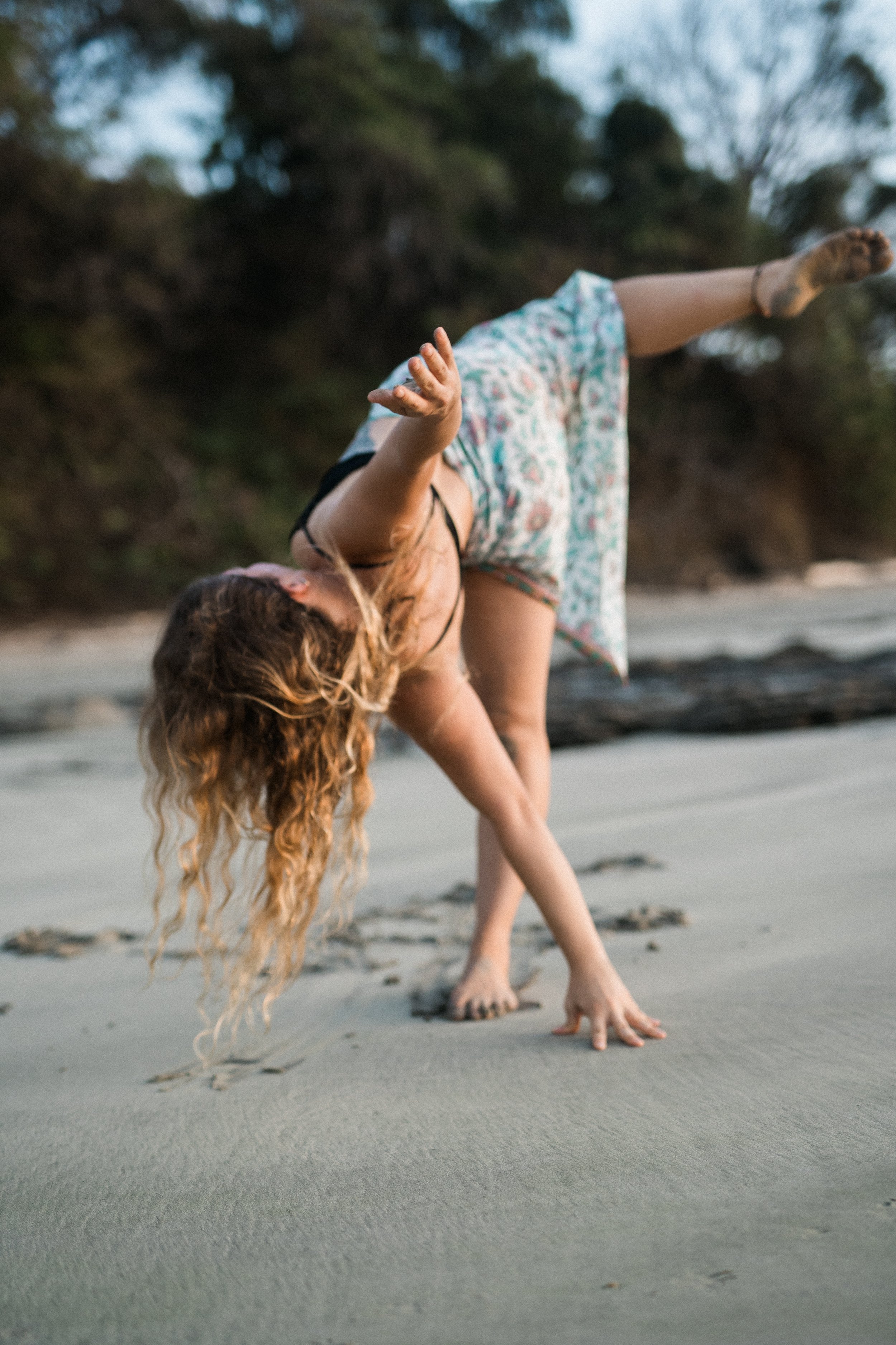 A woman with curly hair doing a handstand on the beach with one leg raised and her head near the sand.