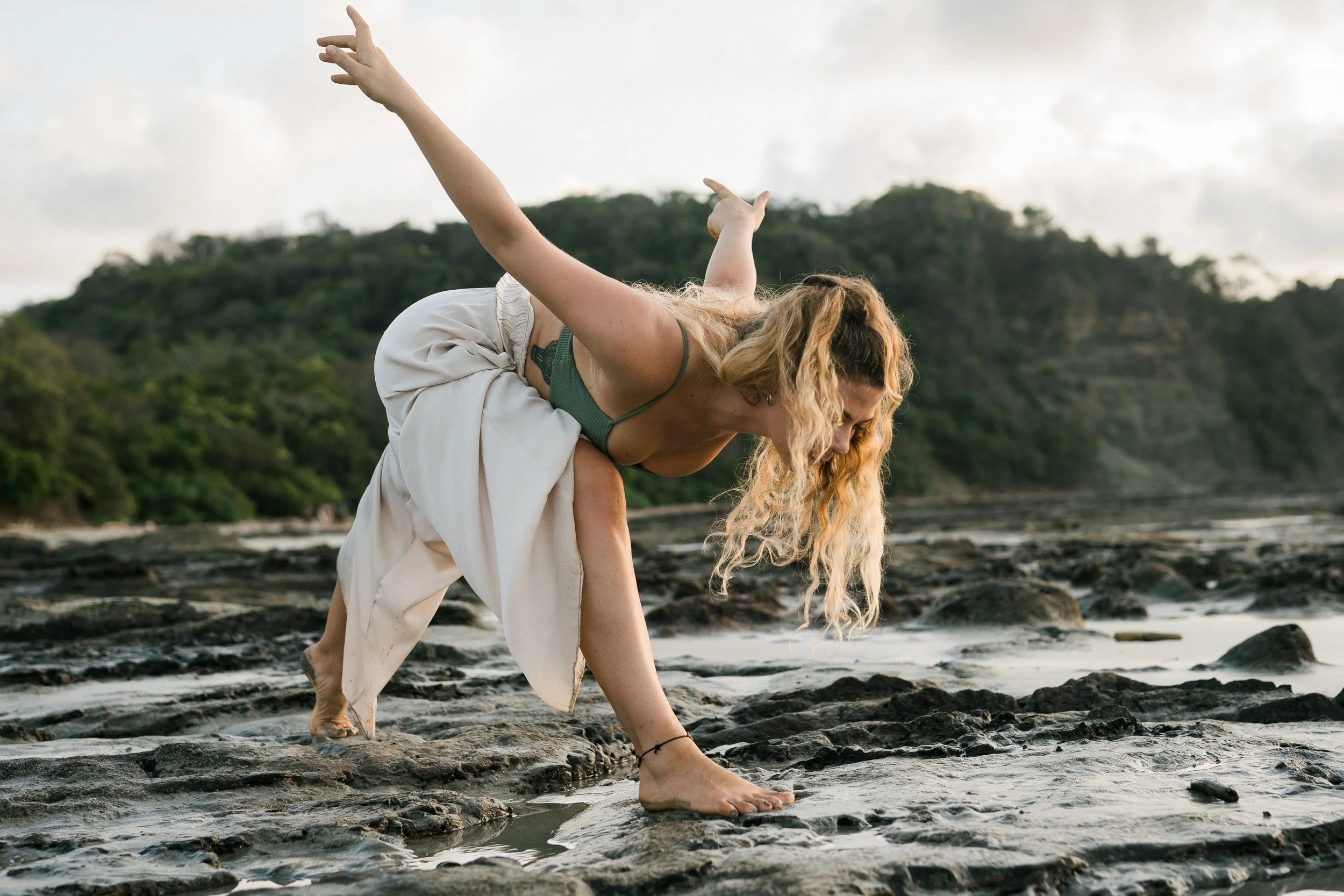 A woman with long, curly blonde hair in a ponytail, wearing a green sports bra and loose white pants, balancing on a rocky shoreline with one hand on the ground and the other extended behind her, against a background of green hills and cloudy sky.