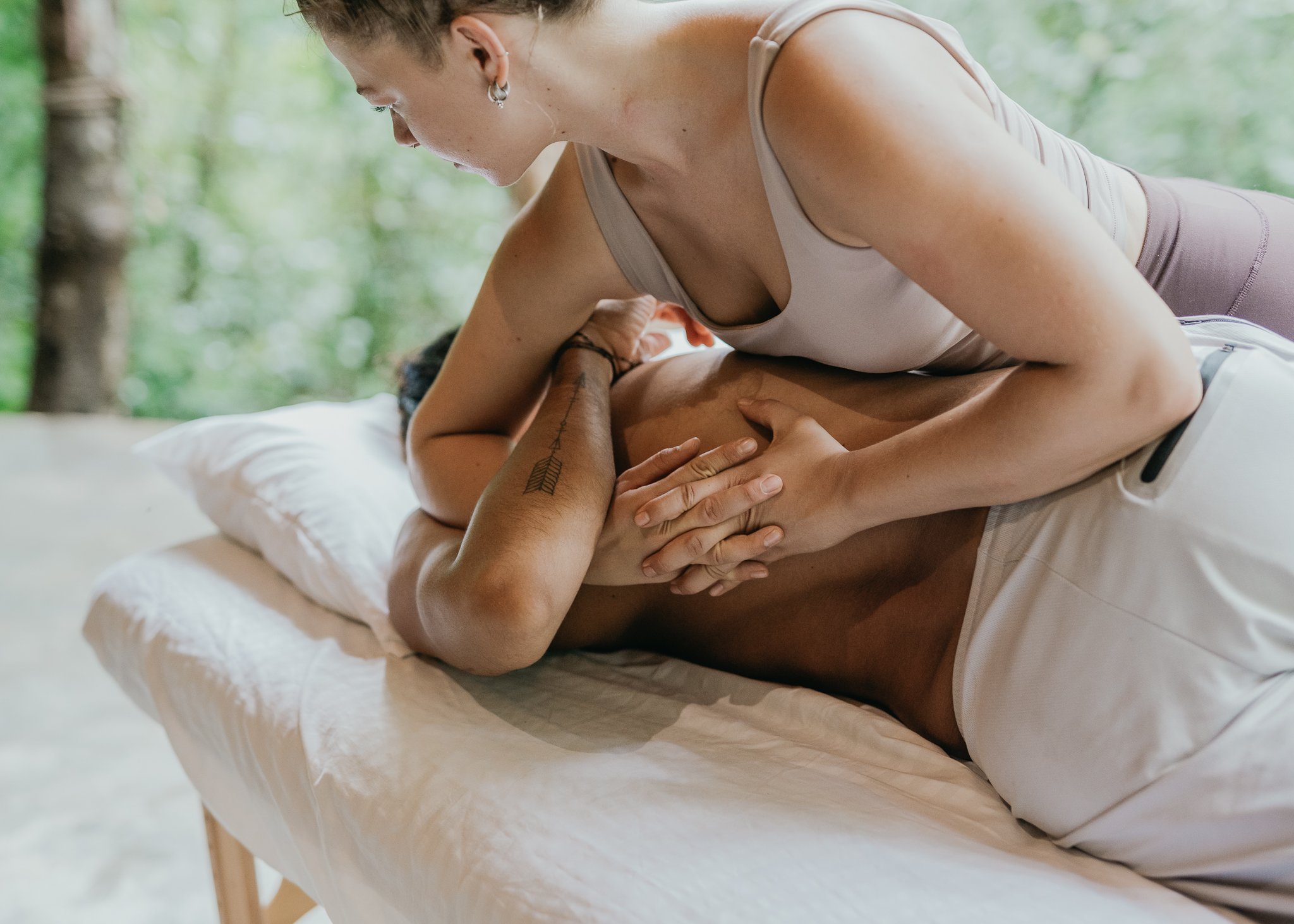 A woman providing a massage to a man lying on a massage table outdoors with a forest background.