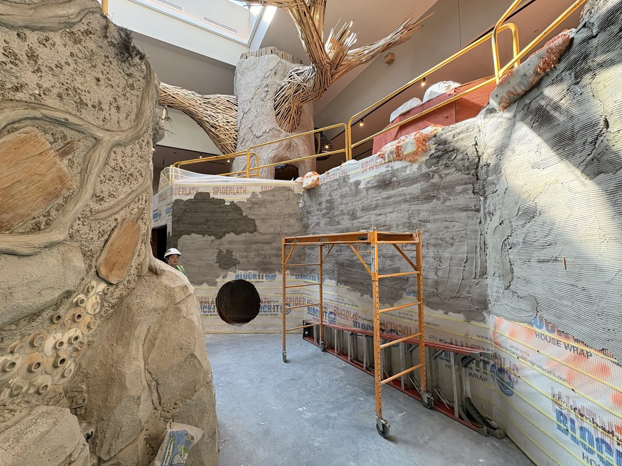 Indoor playground with a large structure resembling a tree, featuring slides and climbing areas, surrounded by children and adults. The ceiling has hanging green light fixtures resembling vines.