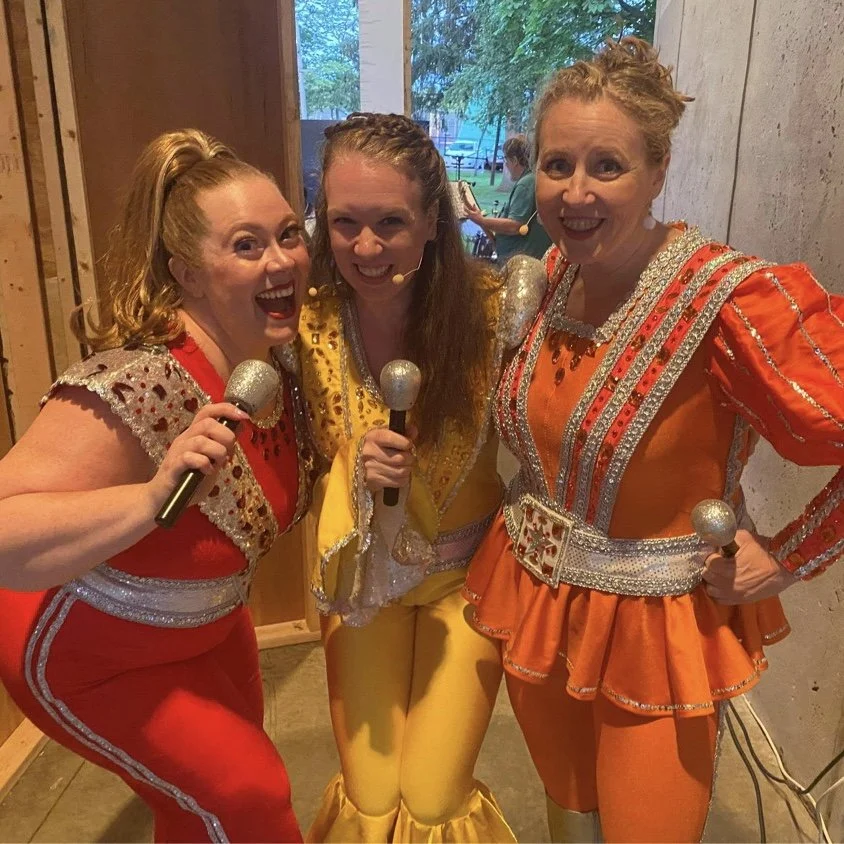 Noddea and two castmates posing backstage in sparkly red, yellow, and orange performance costumes, mid-song.
