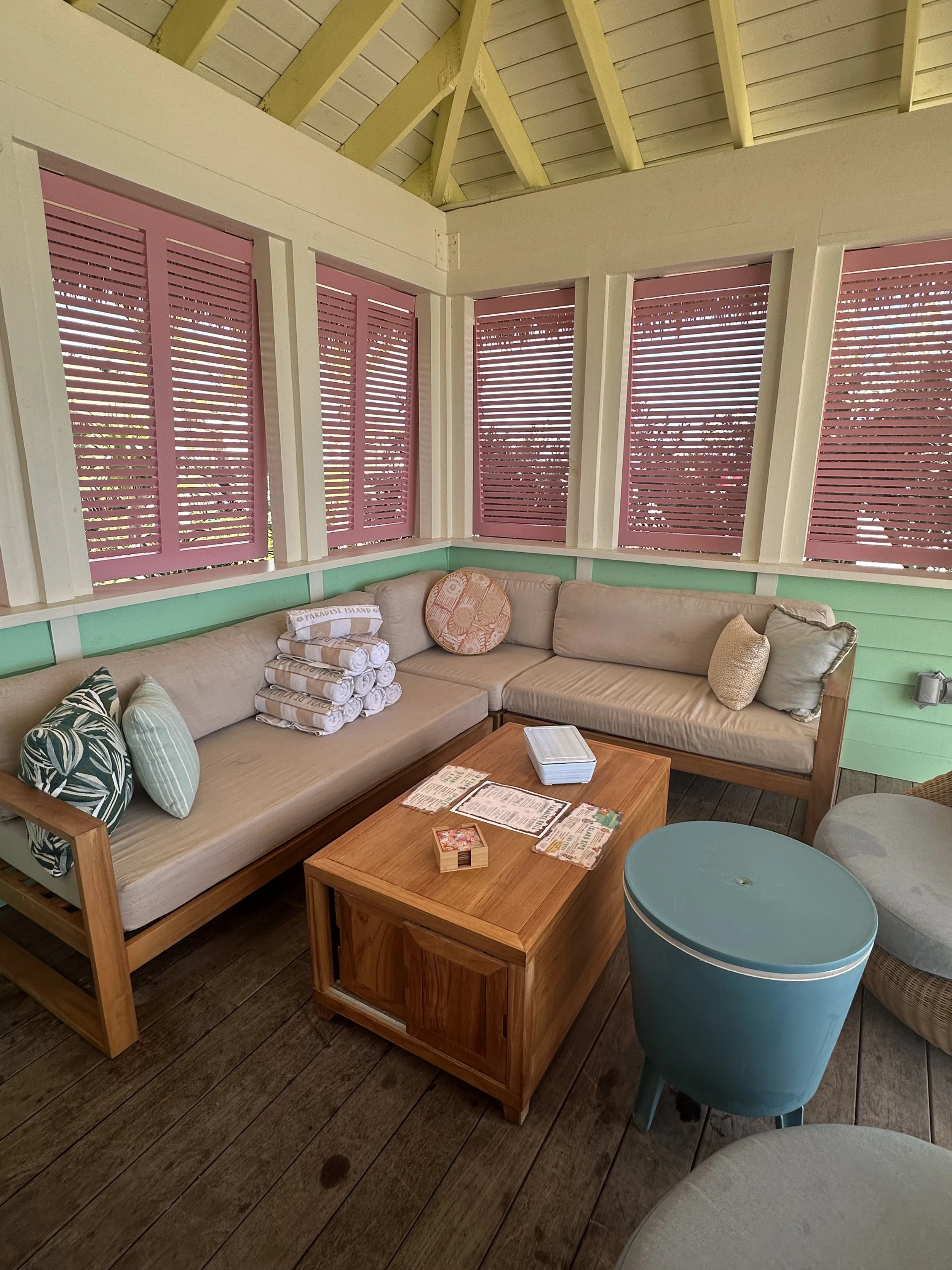 Covered cabana seating area at Royal Beach Club with a cushioned sectional, rolled towels, and pastel green and pink trim.