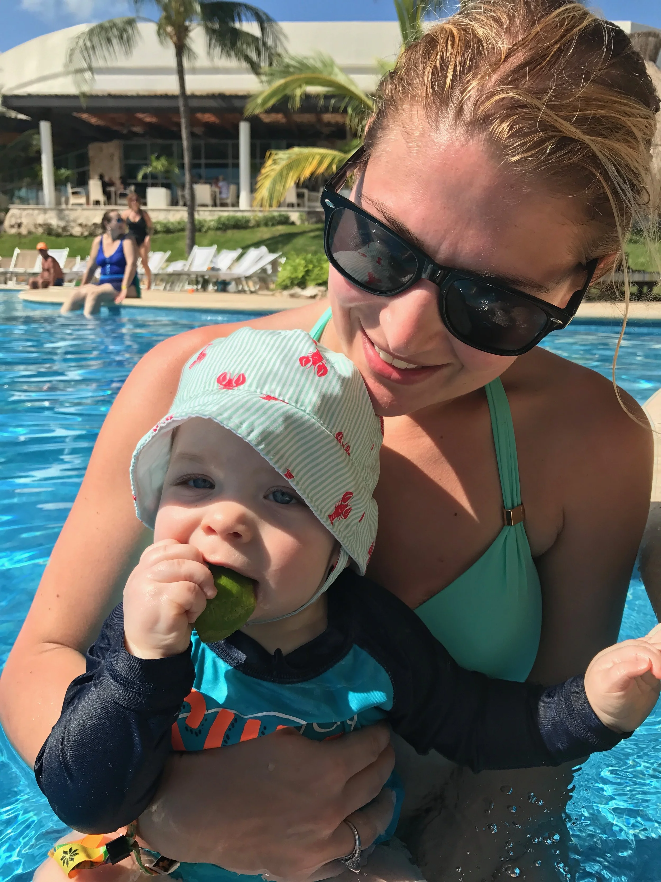 Mom in sunglasses holding a baby in a striped sun hat in a resort pool, palm trees and lounge chairs in the background.