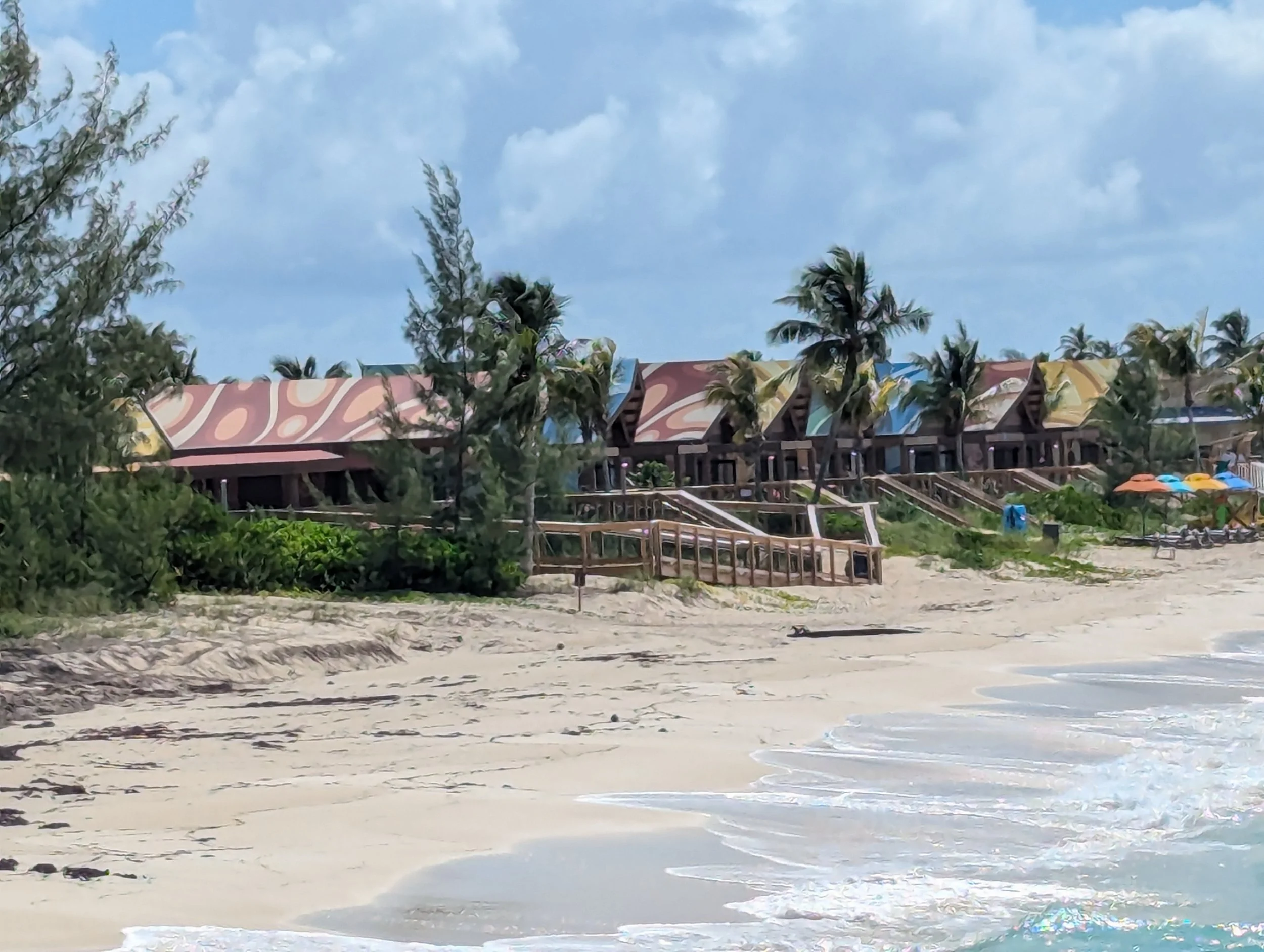 A quiet stretch of beach lined with palm trees and colorful thatched-roof cabana buildings, with gentle waves rolling in along the shore.