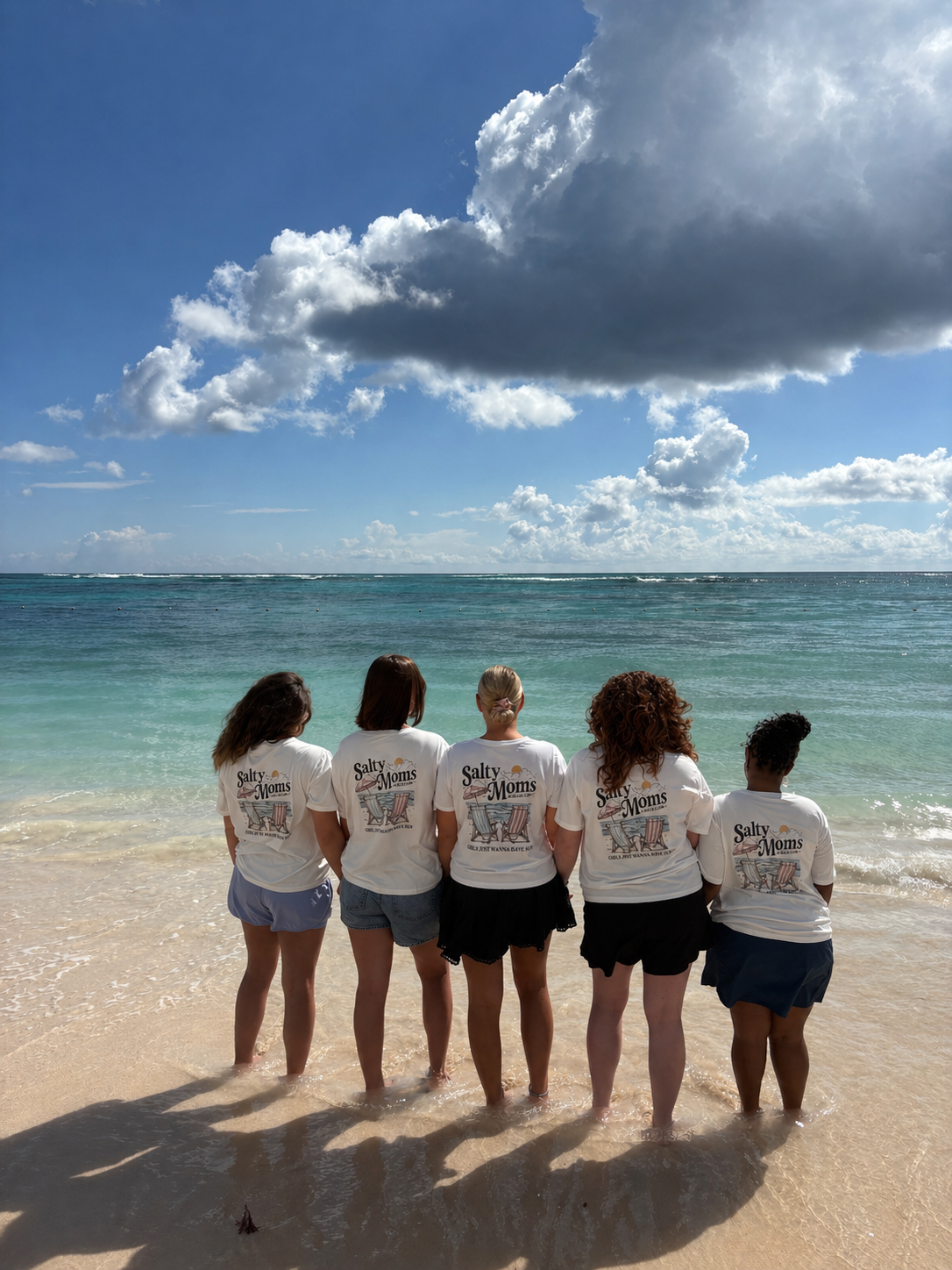 PNG Five women in matching "Salty Moms" t-shirts standing in the surf, backs to camera, looking out at turquoise ocean water.