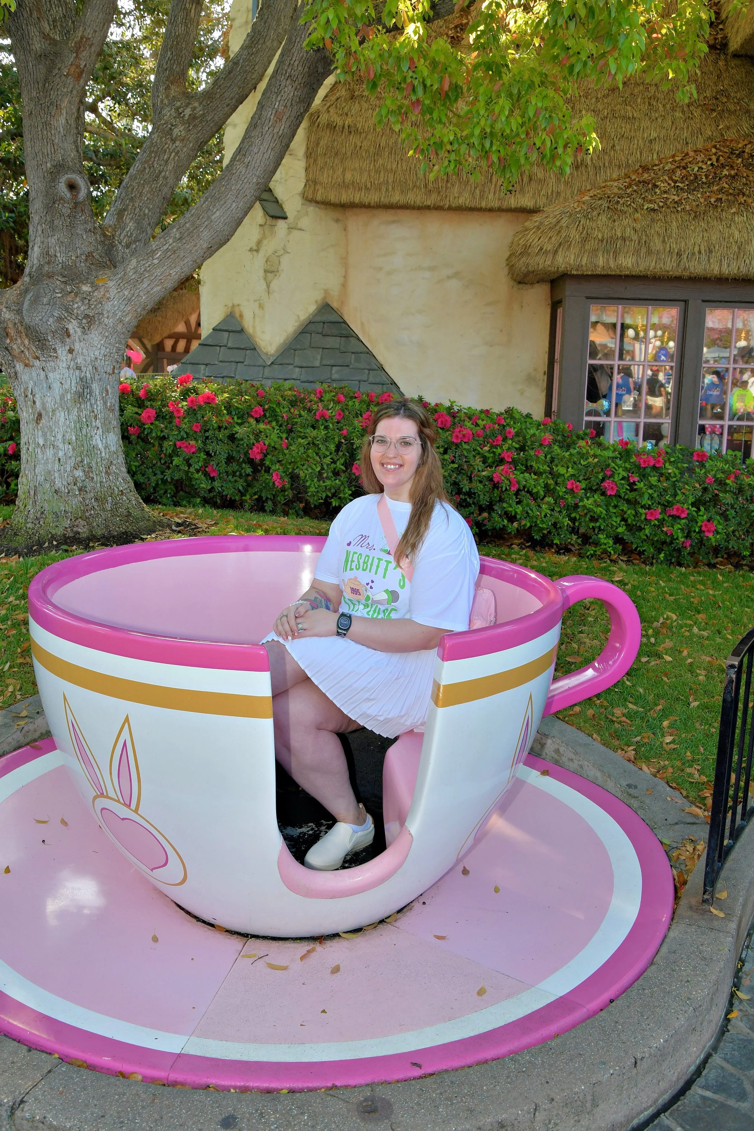Jess smiling in a giant pink teacup photo spot at Disneyland, surrounded by blooming pink rose bushes and a thatched-roof building.