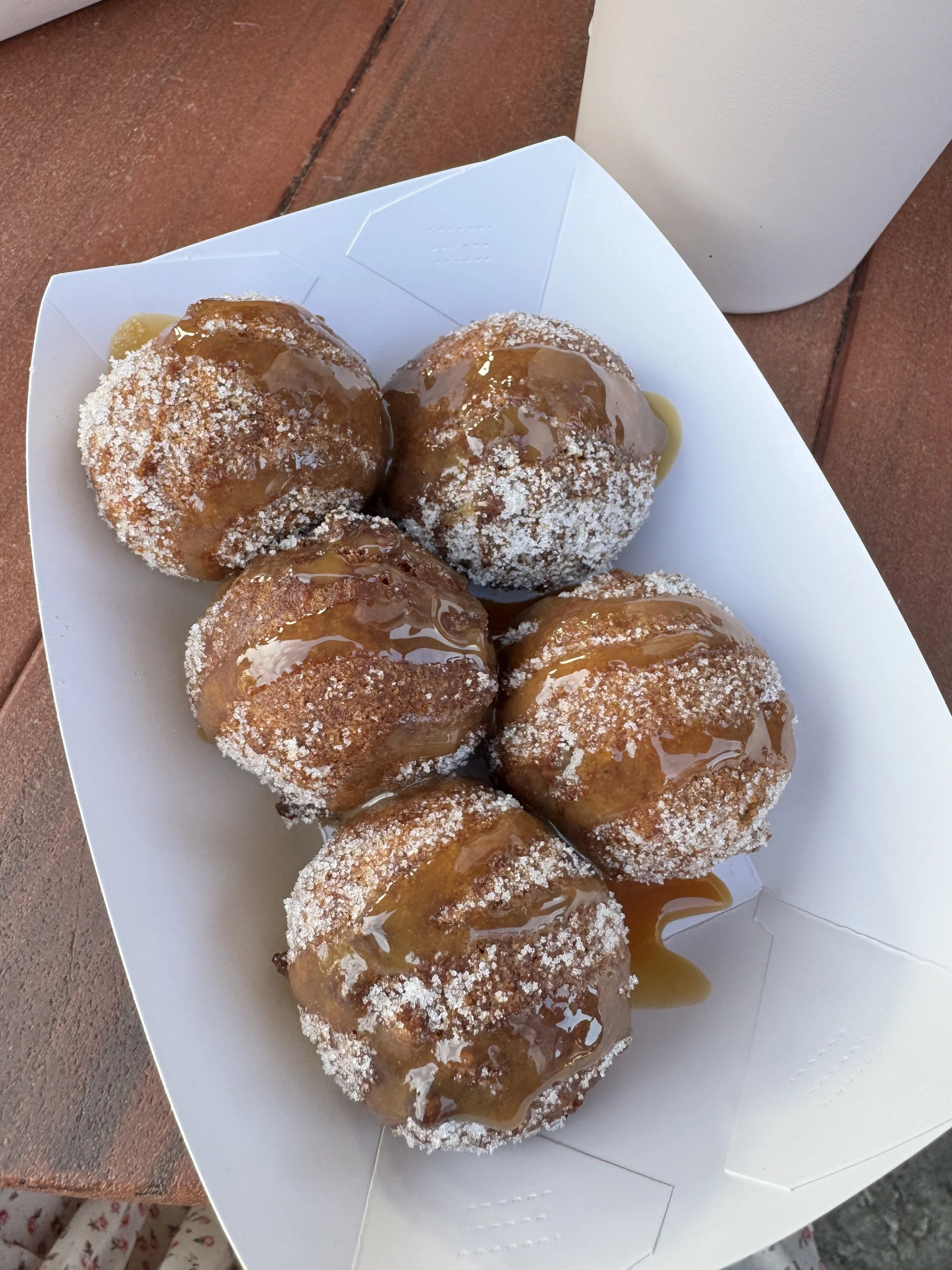 Five sugar-dusted fried dough bites drizzled with caramel sauce, served in a paper festival tray on a wooden table.