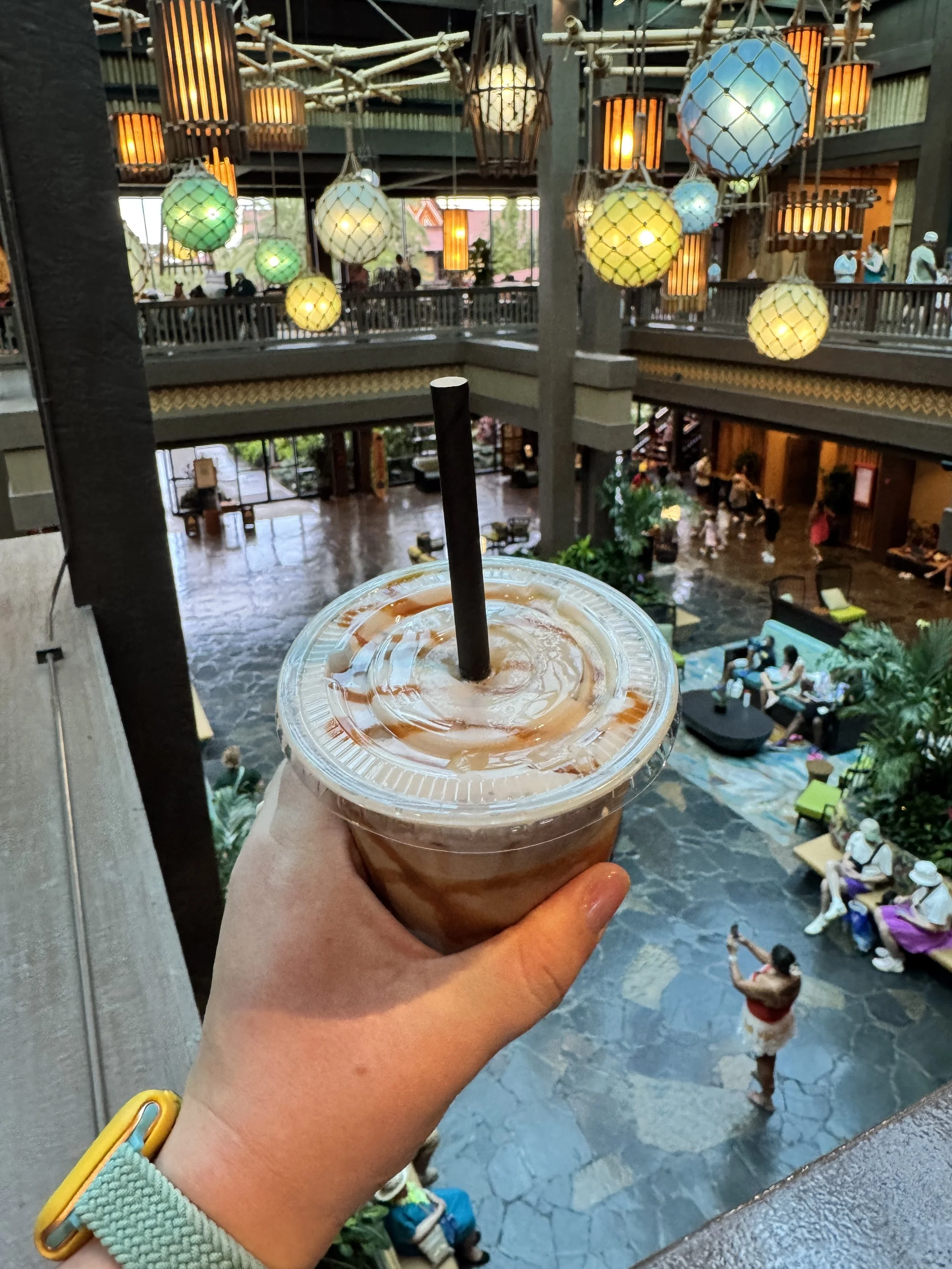 A hand holds a caramel iced coffee above the open, multi-story lobby of Disney's Polynesian Village Resort, with woven lanterns and glass fishnet floats overhead.