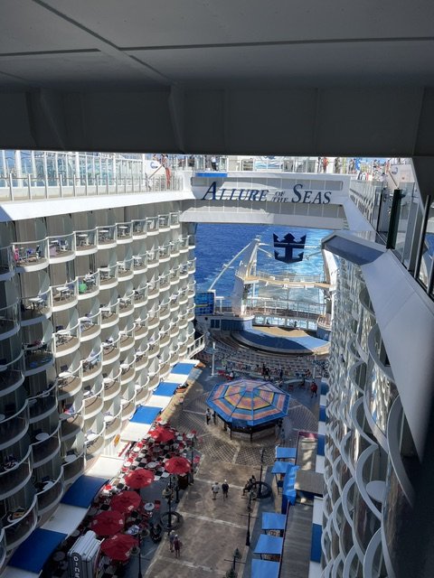 View down into the open-air Boardwalk neighborhood aboard Allure of the Seas, lined with balconies, umbrellas, and the ship's name overhead.