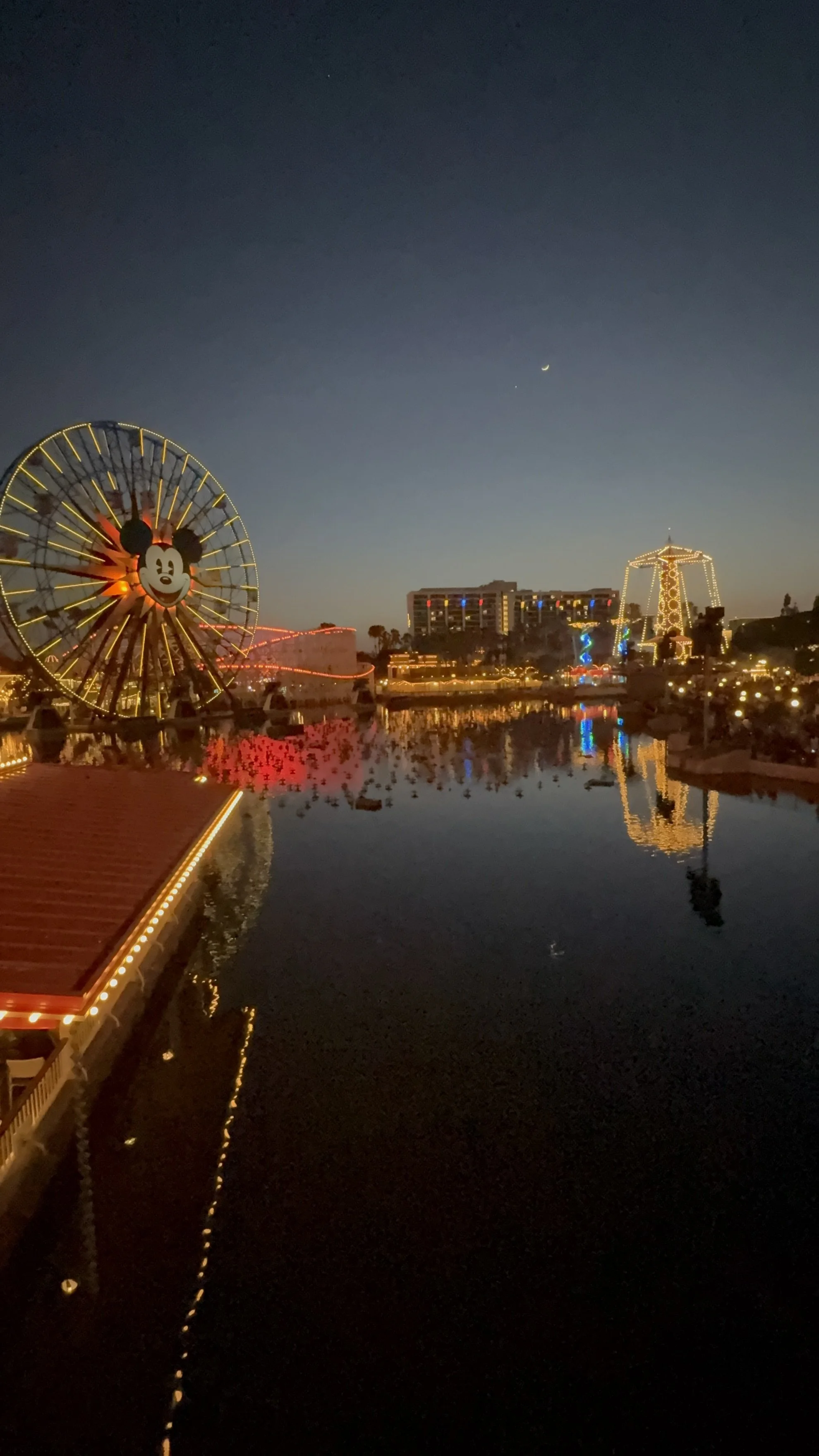 a small body of water with a ferris wheel with Mickey Mouse face on the left and hotel on the right