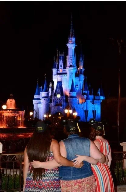 Three friends with their arms around each other, looking toward Cinderella Castle lit blue at night in Magic Kingdom.