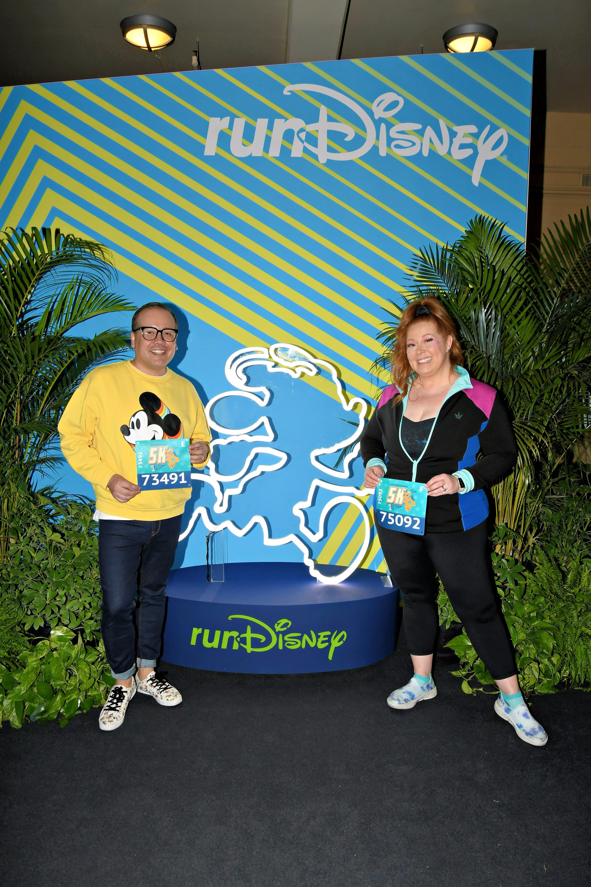 Noddea and her husband posing with their runDisney 5K race bibs in front of the runDisney photo backdrop.