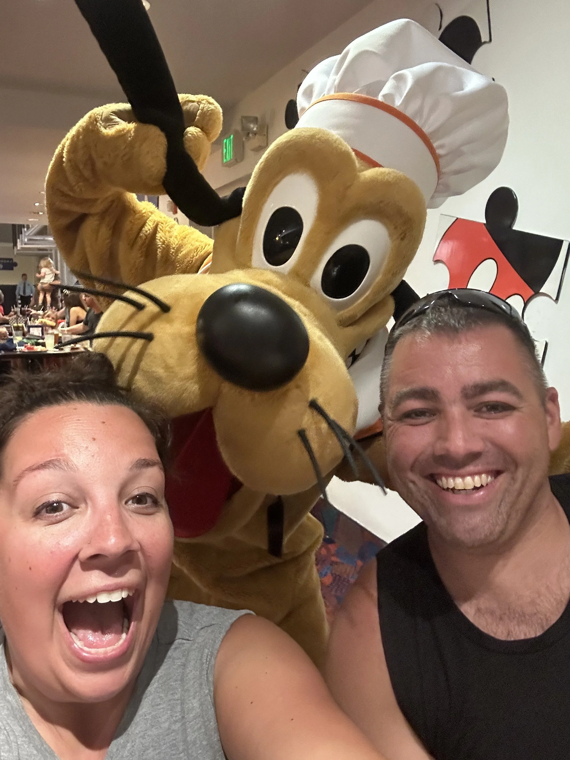 Darcy Roy and her husband posing with Pluto in his chef's hat at a Disney character dining experience.