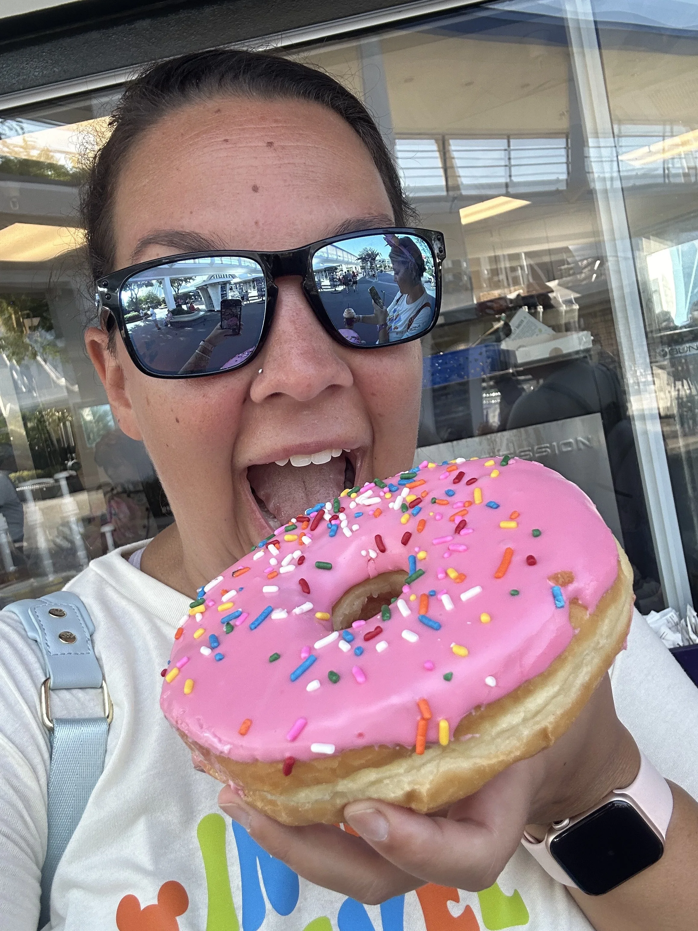 Travel advisor Darcy Roy smiling behind an oversized pink sprinkle donut outside a Walt Disney World snack stand.