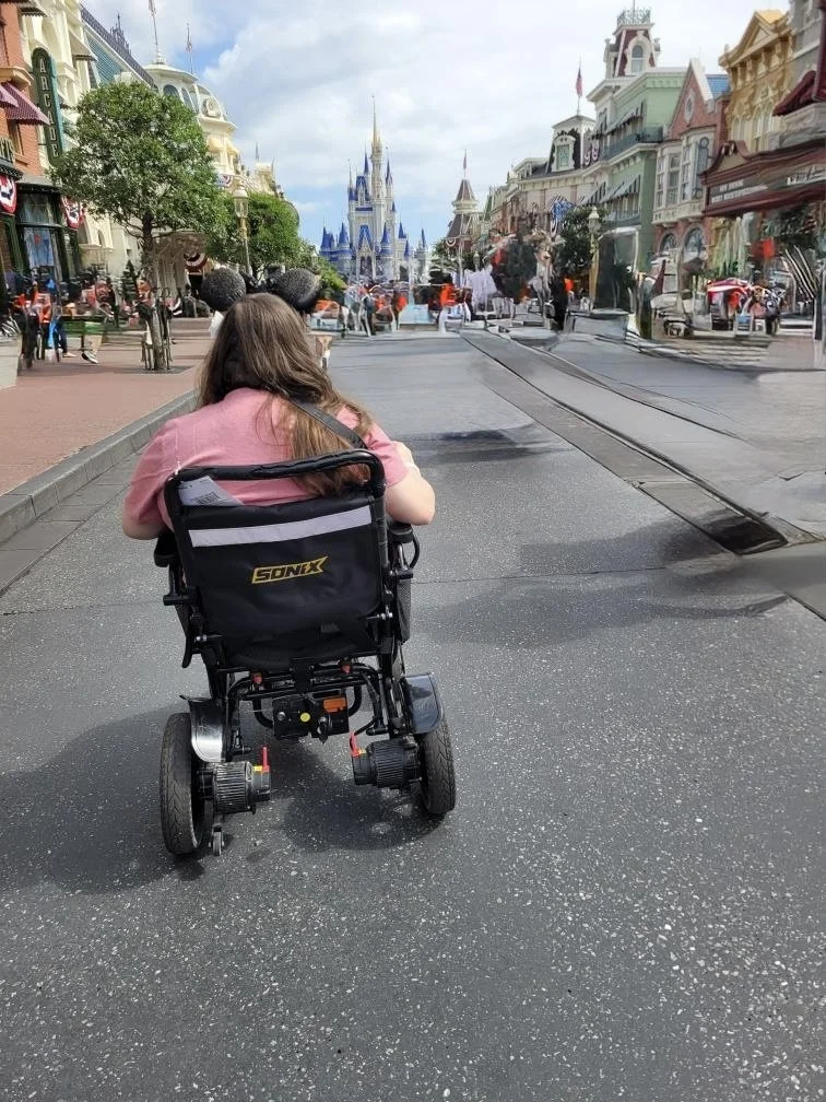Becca, seen from behind in her power wheelchair, riding down Main Street U.S.A. toward Cinderella Castle at Magic Kingdom.