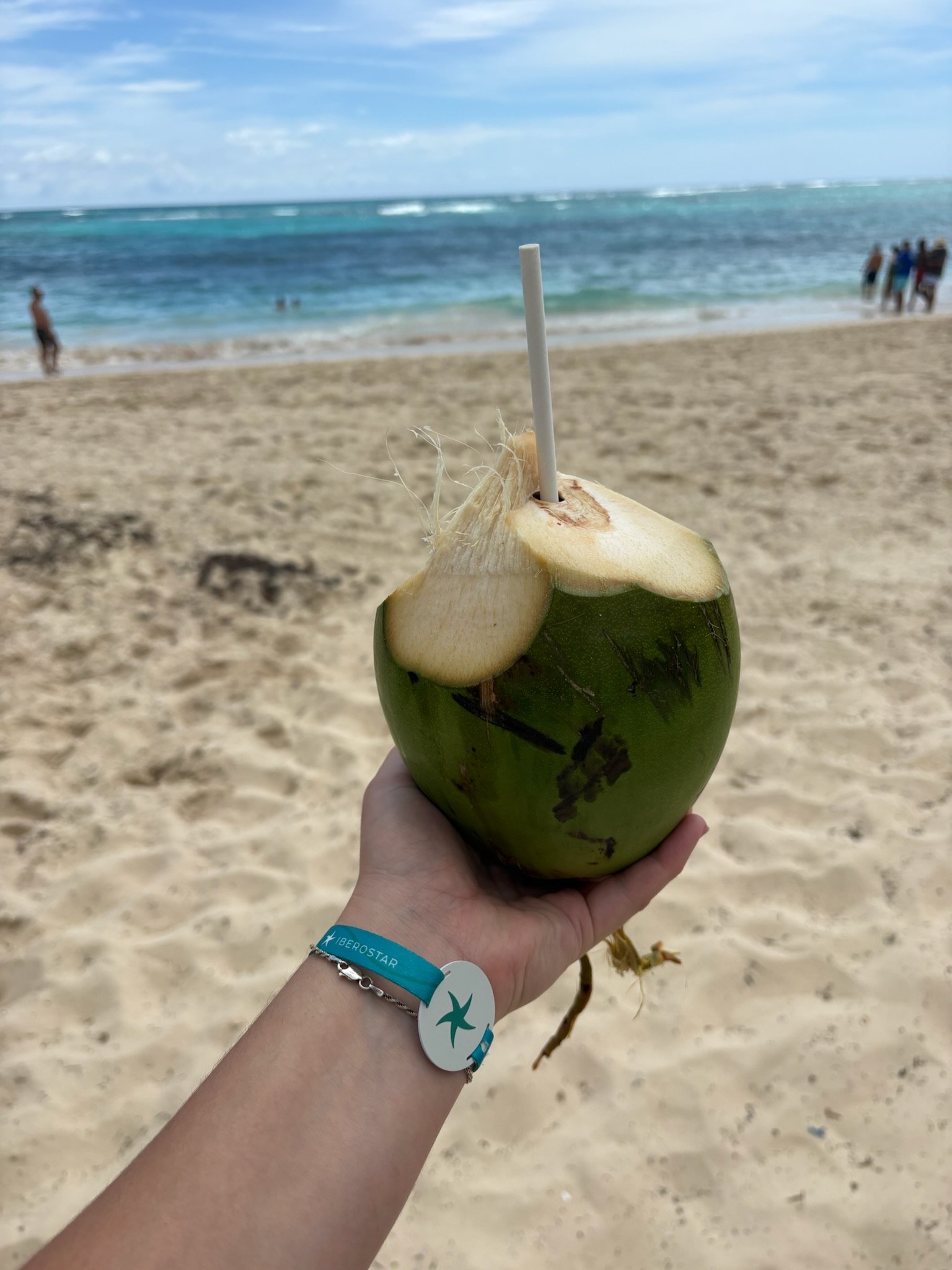 a hand holding out a coconut cut open with a straw in it with a beach view behind it