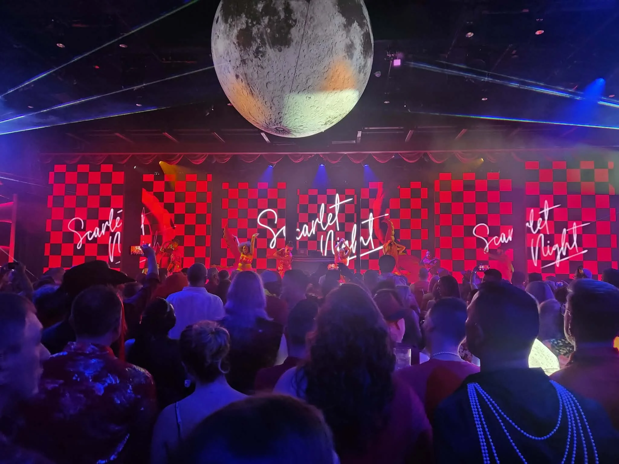 crowd of people in a dark room with a large red checkered screen that says scarlet night