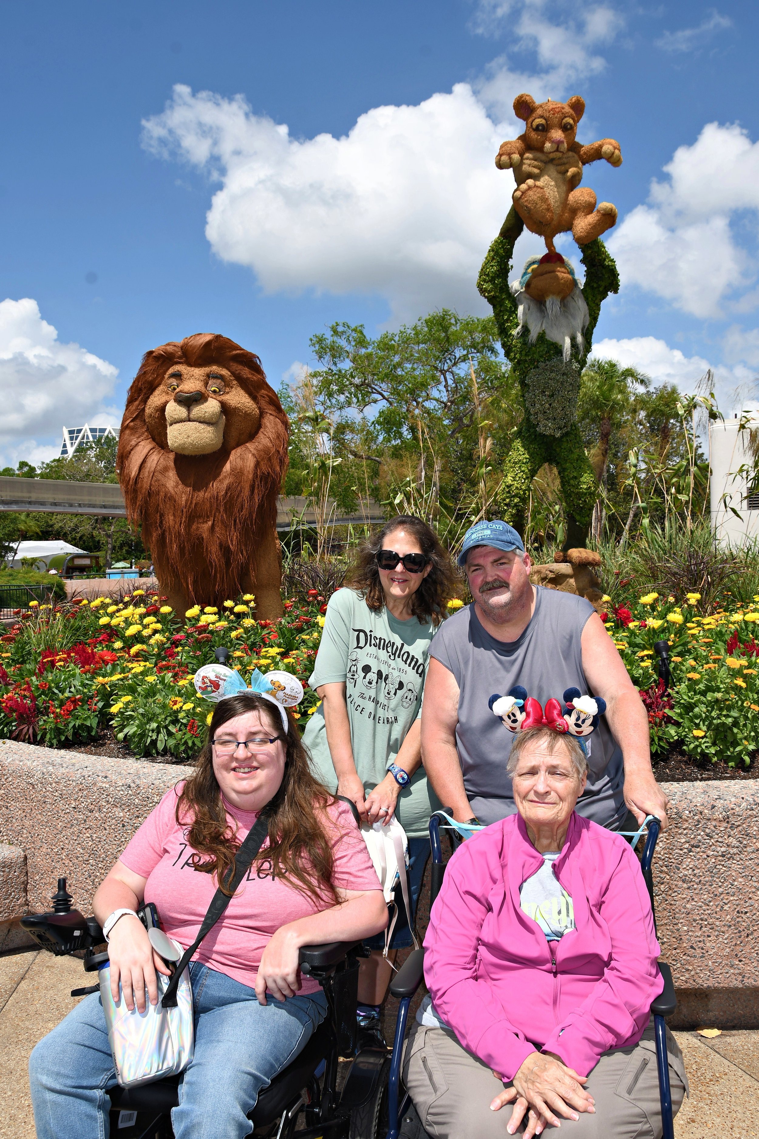 A family of four, including two people seated in wheelchairs, pose together in front of large Lion King-themed topiaries (Mufasa and Rafiki holding baby Simba) surrounded by red and yellow flowers.