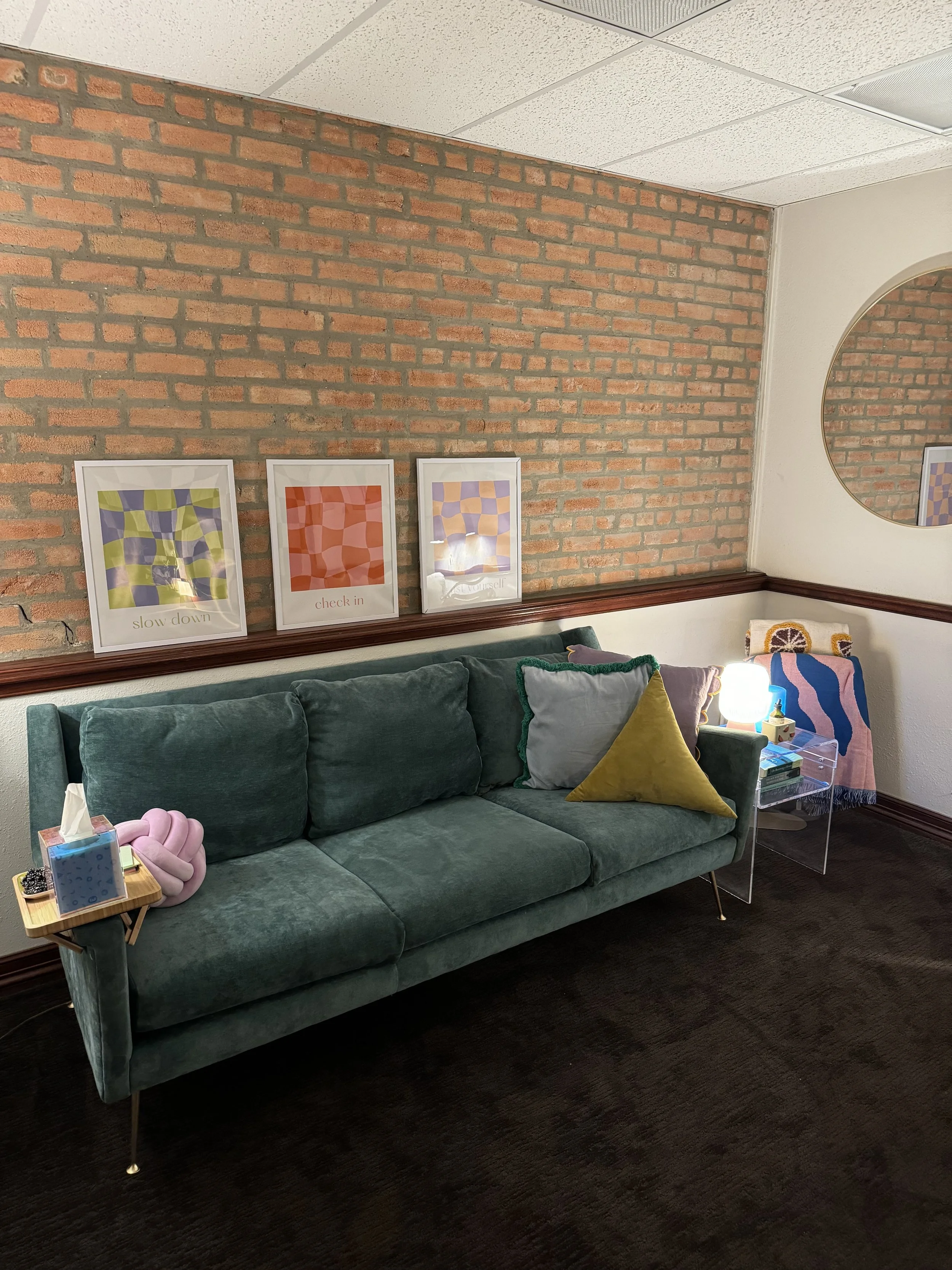 Living room with a green velvet sofa, decorative pillows, a small side table with tissues, framed abstract art prints on brick wall, and a circular mirror on white wall.