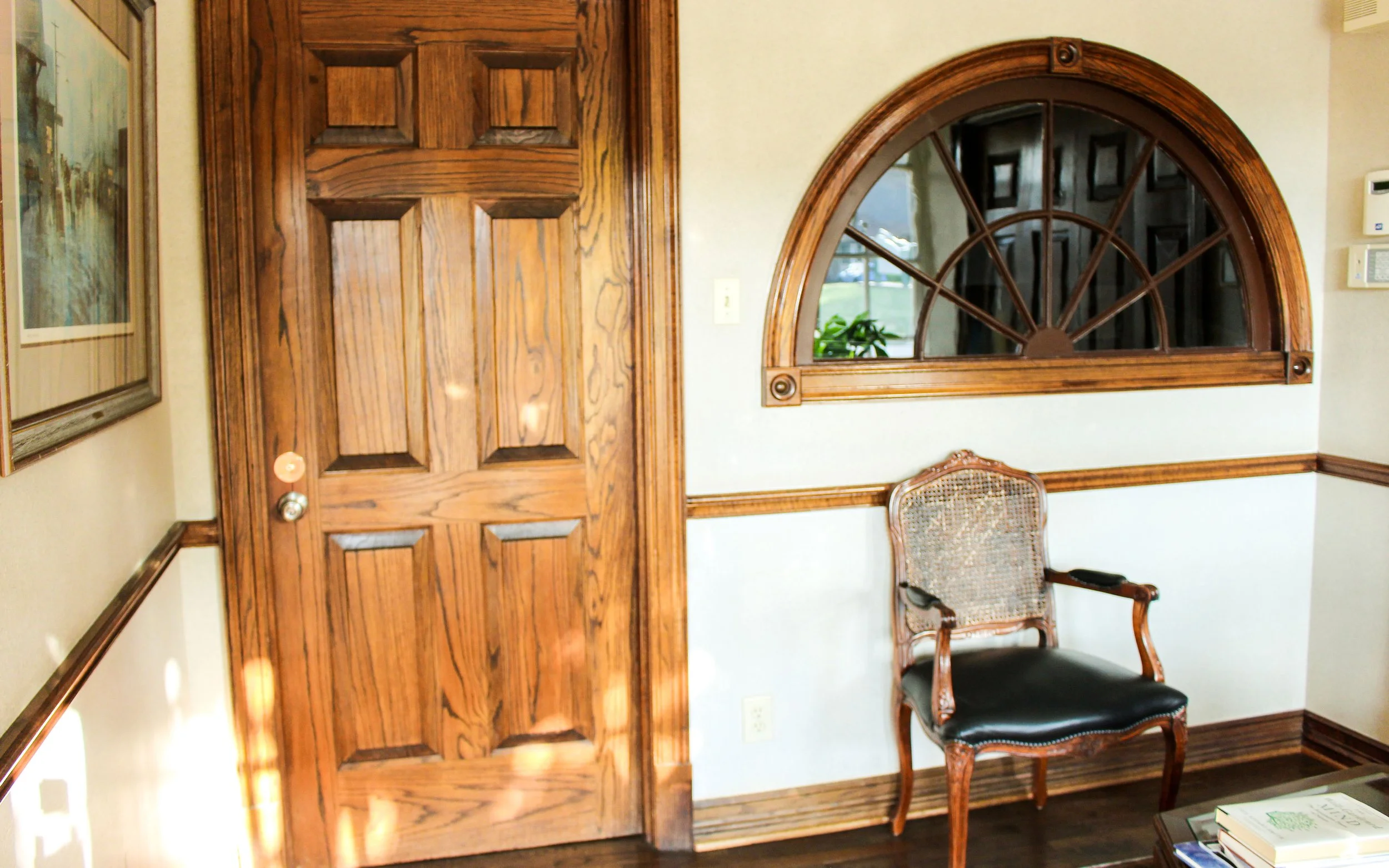 Interior of a room with a wooden door, a decorative window, a vintage armchair with a cane back and black seat cushion, a framed picture on the wall, and a table with books.