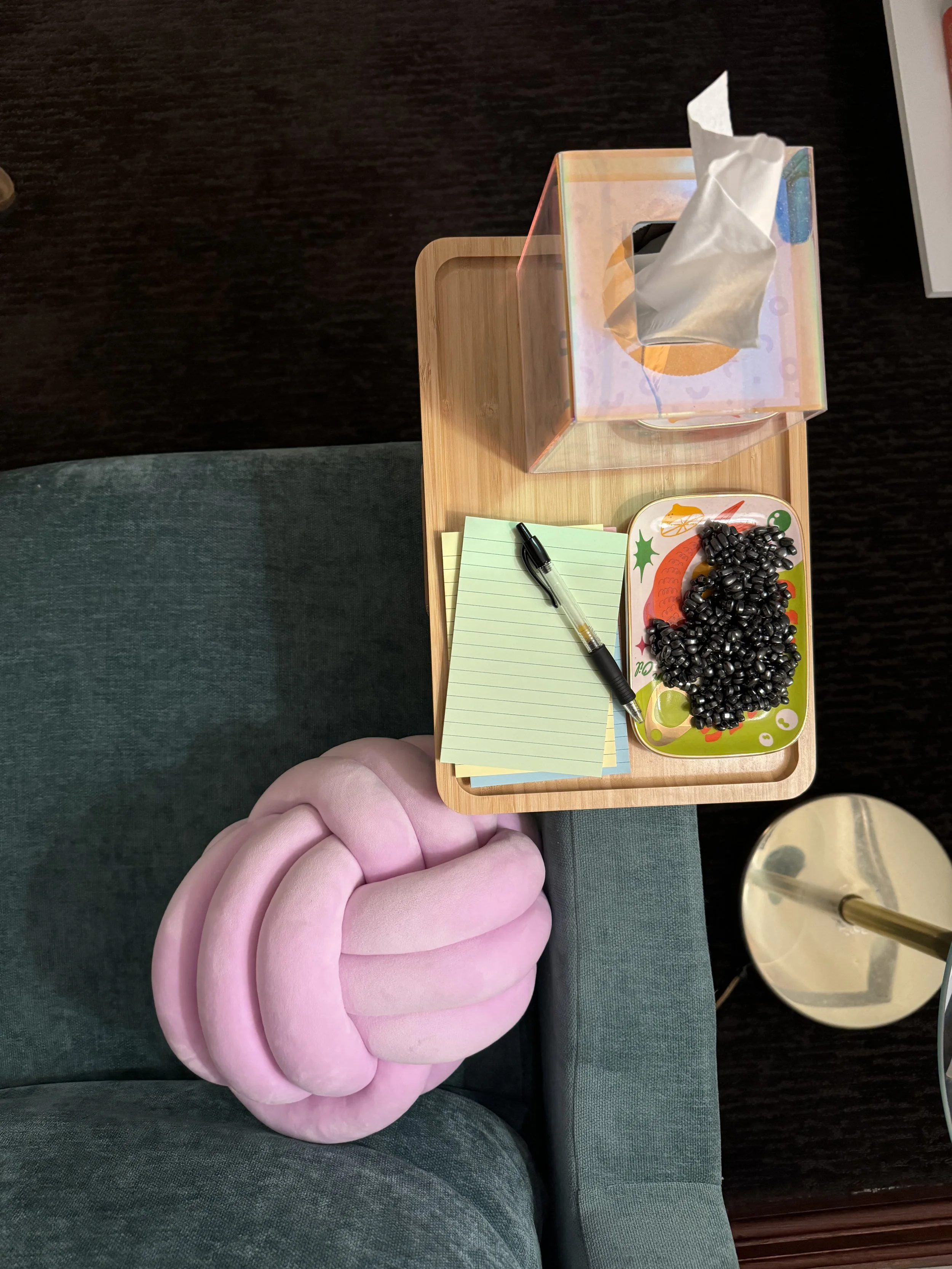 A pink, knot-shaped cushion on a green sofa. On a wooden side table, there are yellow sticky notes, a black pen, a decorative plate with black candies, a tissue box, and a lamp with a gold stand.