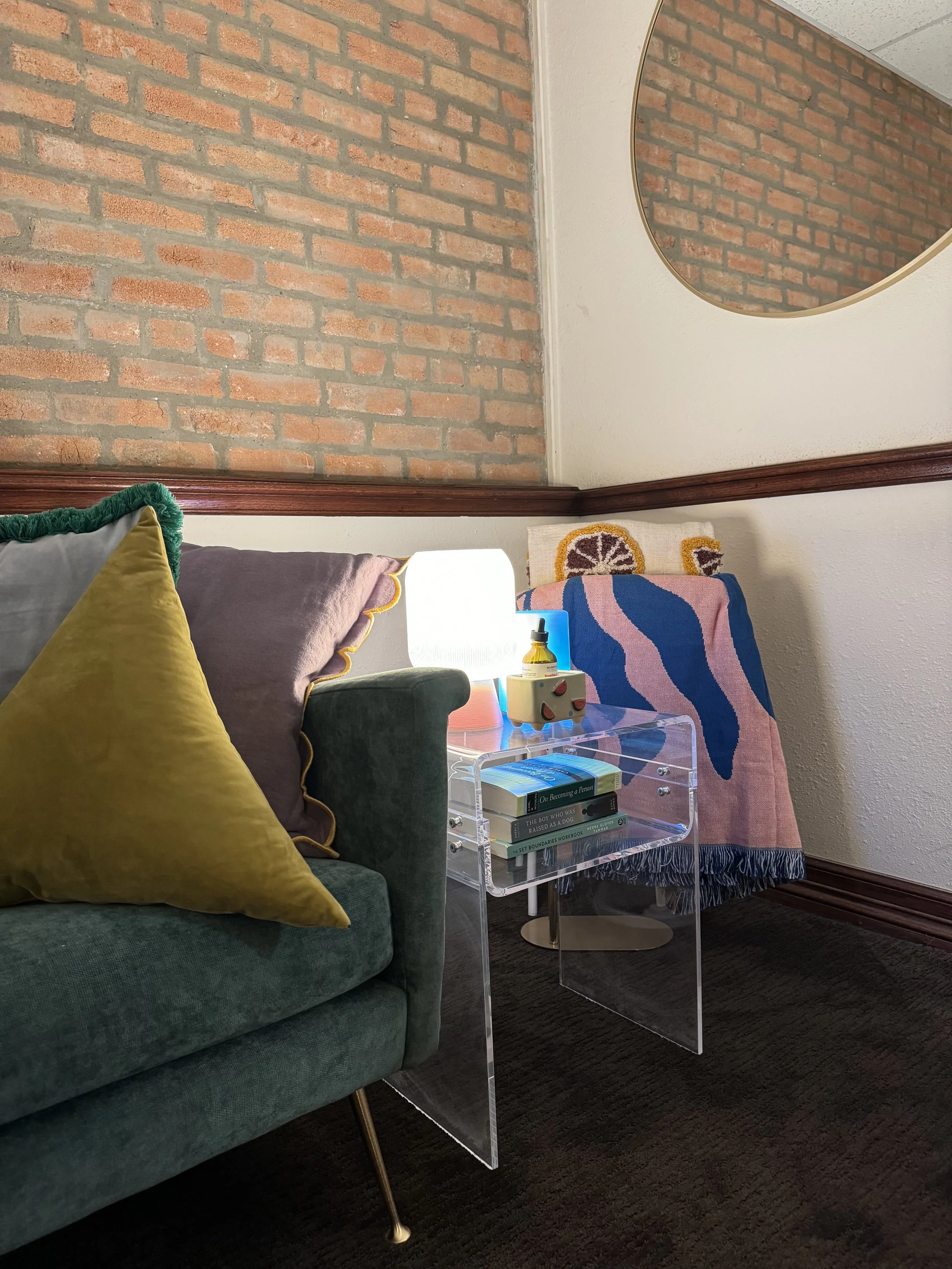 A cozy living room corner featuring a green velvet sofa with a yellow and a lilac pillow, a clear acrylic side table with books, a modern lamp, and decorative items, against a red brick wall and white wall with a round mirror.