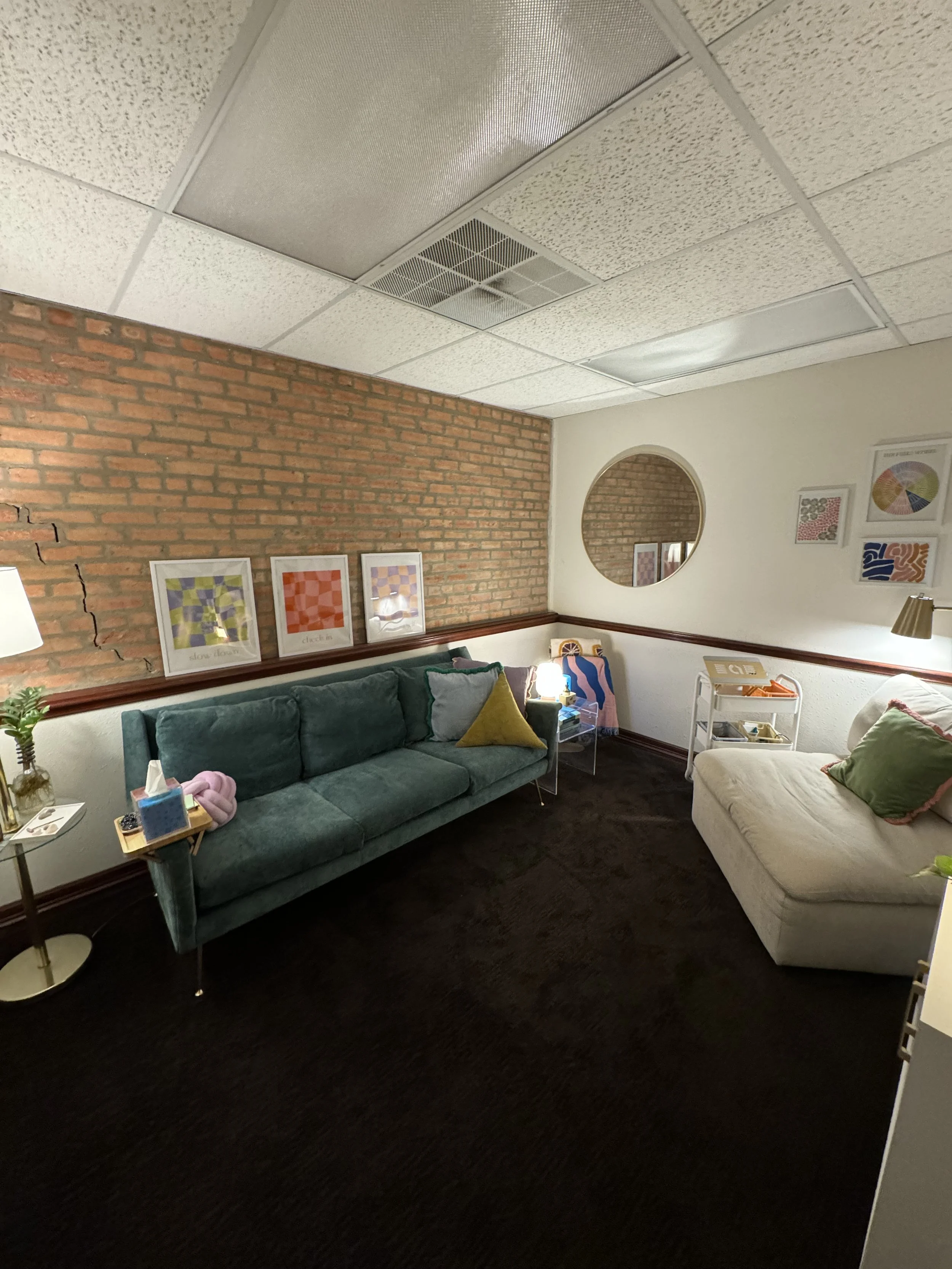 A cozy living room with a brick accent wall, colorful abstract framed artwork, a green velvet sofa with pillows, and a beige chaise lounge, with a small side table, decorative plants, and framed art on the walls.