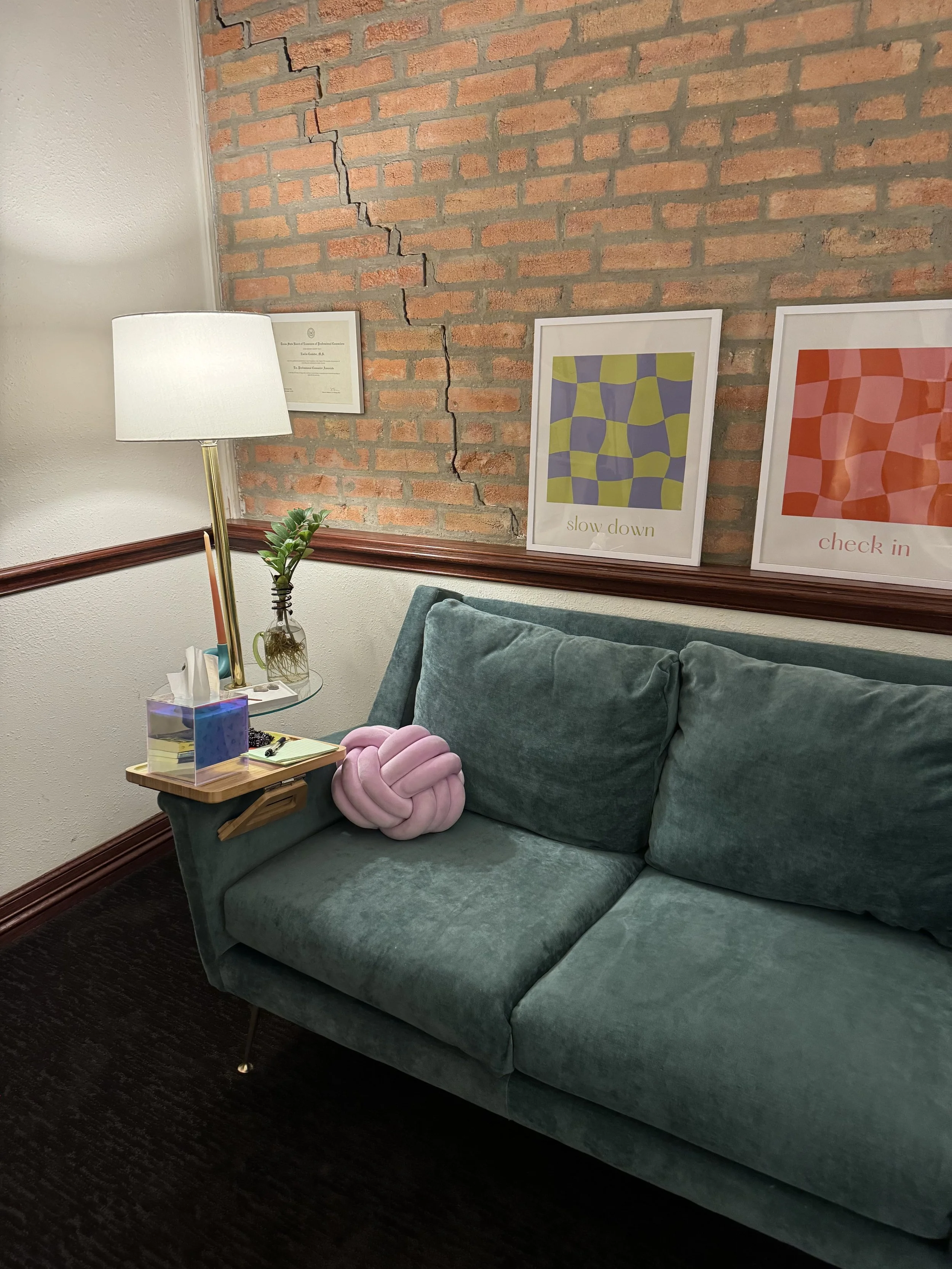 A teal sofa with a pink knotted knot pillow, a small side table with a tissue box, a lamp, and decorative items, placed against a brick wall with a large crack, and framed artwork with colorful abstract patterns and words.