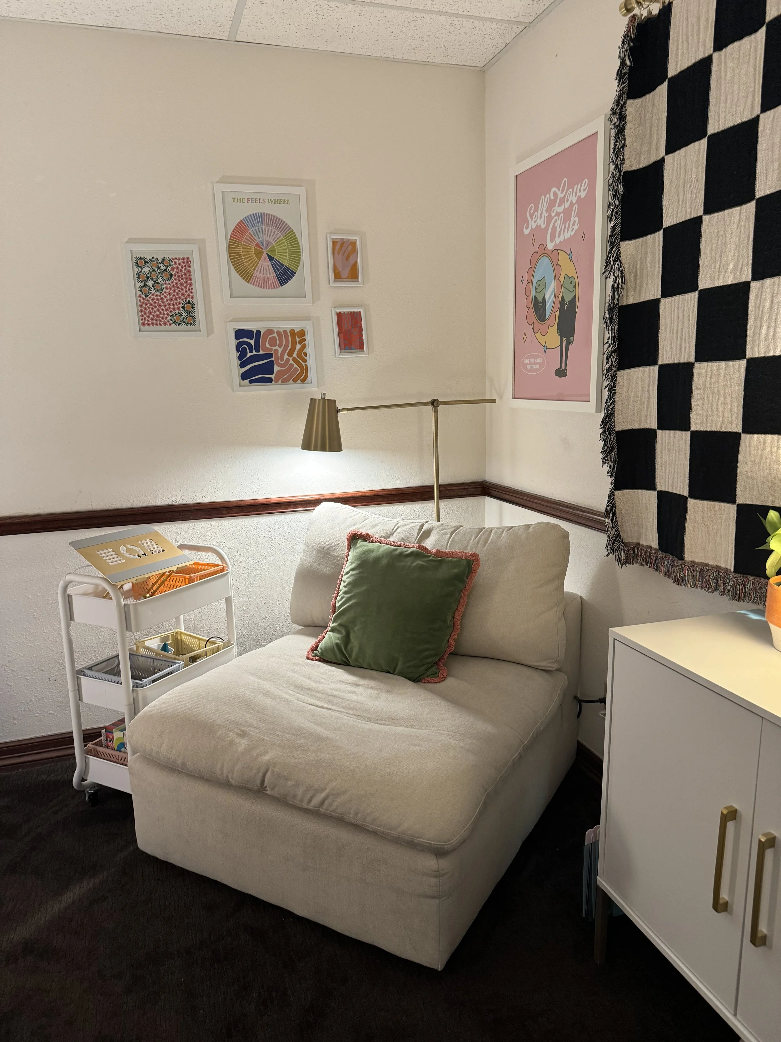 A cozy corner with a white armchair and green pillow, next to a white cabinet, a small rolling cart, and a wall decorated with framed artwork and a large pink poster reading 'Self Love Club.'