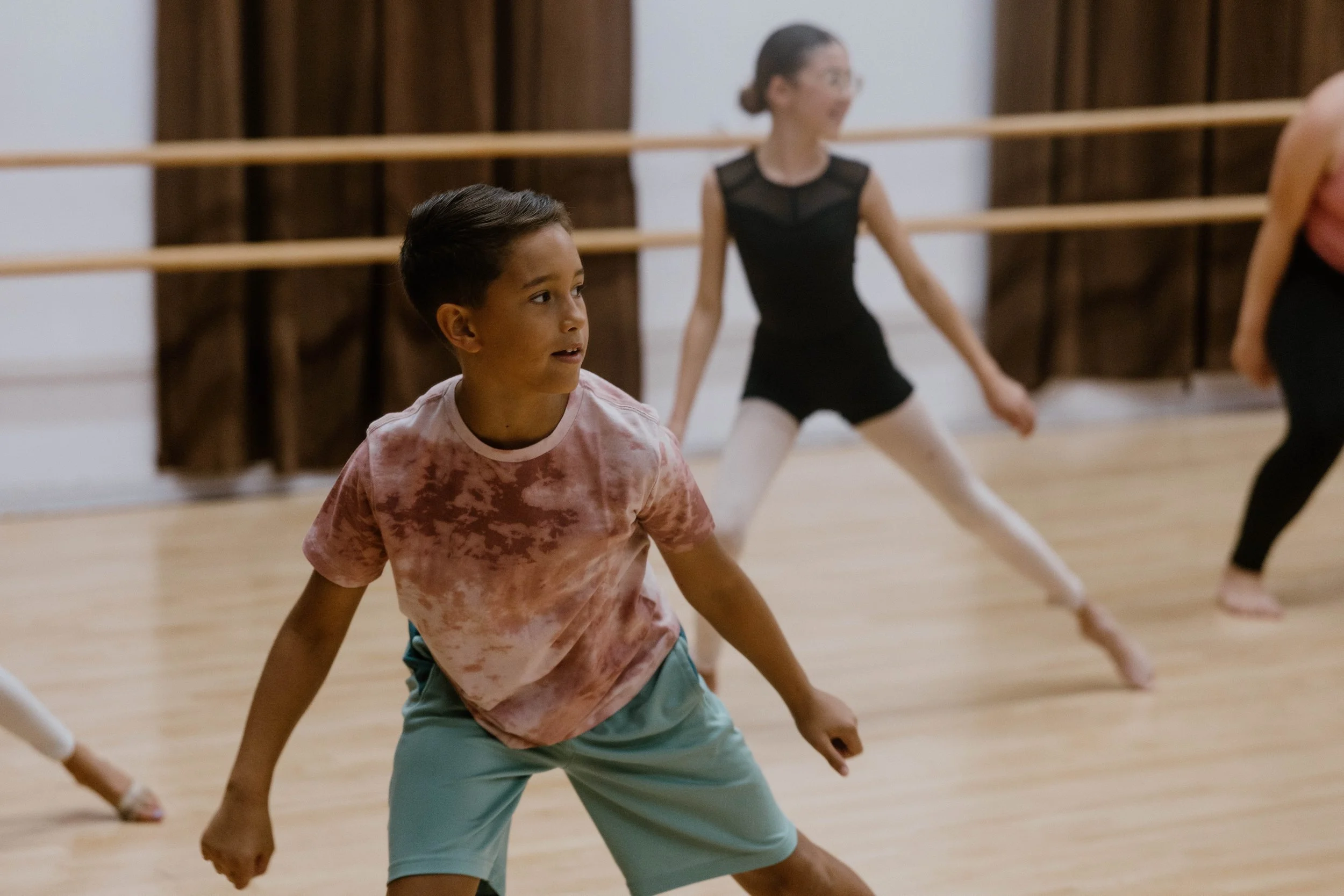 A young RAD dancer mid-movement in class, engaged and focused, with classmates practicing in the background.