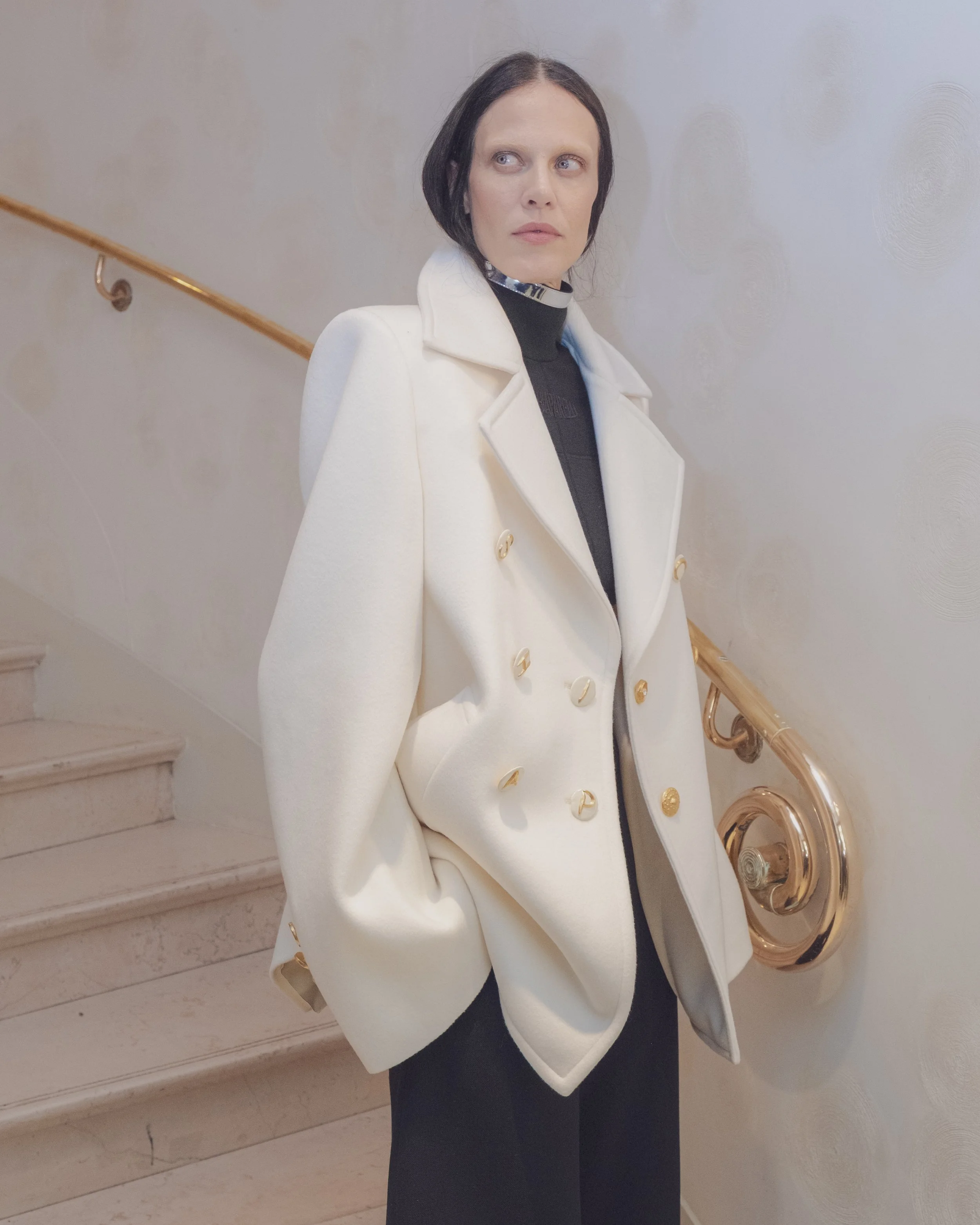 Woman with short dark hair wearing a white coat and black turtleneck, holding a gold musical instrument, standing near a staircase with a gold handrail.