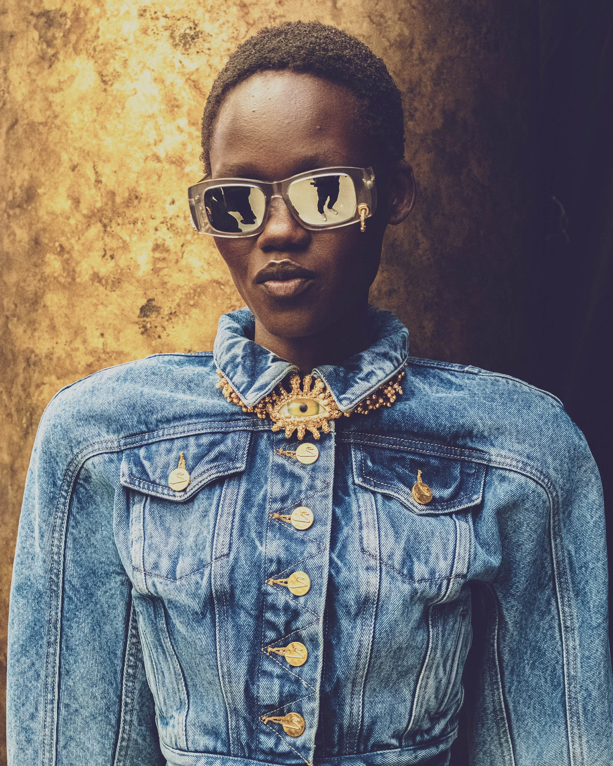 Close-up of a person with short curly hair wearing reflective sunglasses, dark lipstick, a denim jacket with gold buttons, and a statement gold necklace with an eye pendant. The background is a textured brown wall.