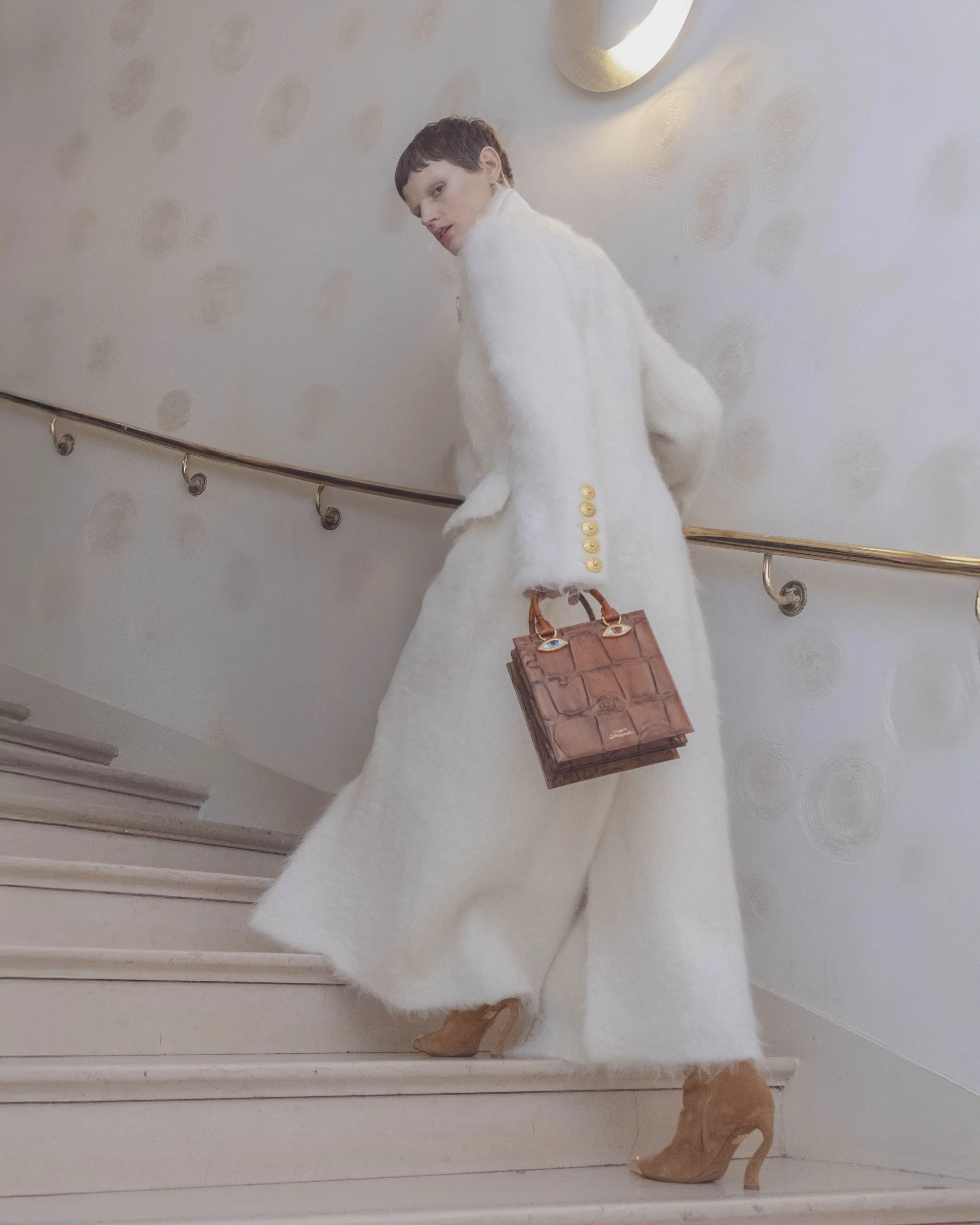 A woman in a white dress with long sleeves, high heels, and a brown handbag, standing on a staircase, looking back over her shoulder. The background features a wall with golden decorative circular accents and a gold circular mirror. The staircase has