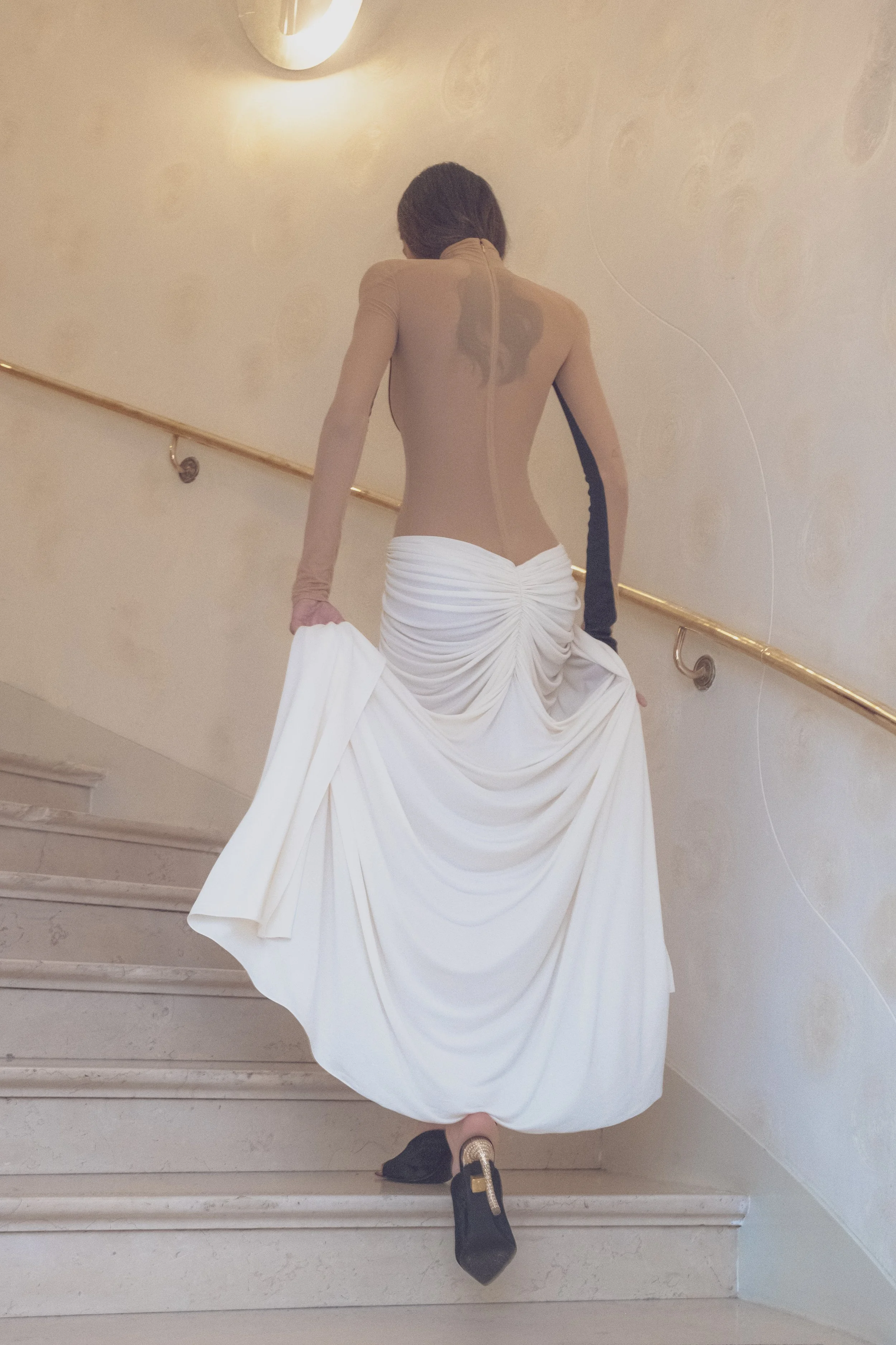 Person ascending stairs, wearing a beige top with a heart-shaped back cutout and a white draped skirt, black and gold shoes, holding a white cloth, with a curved gold railing on a beige wall.