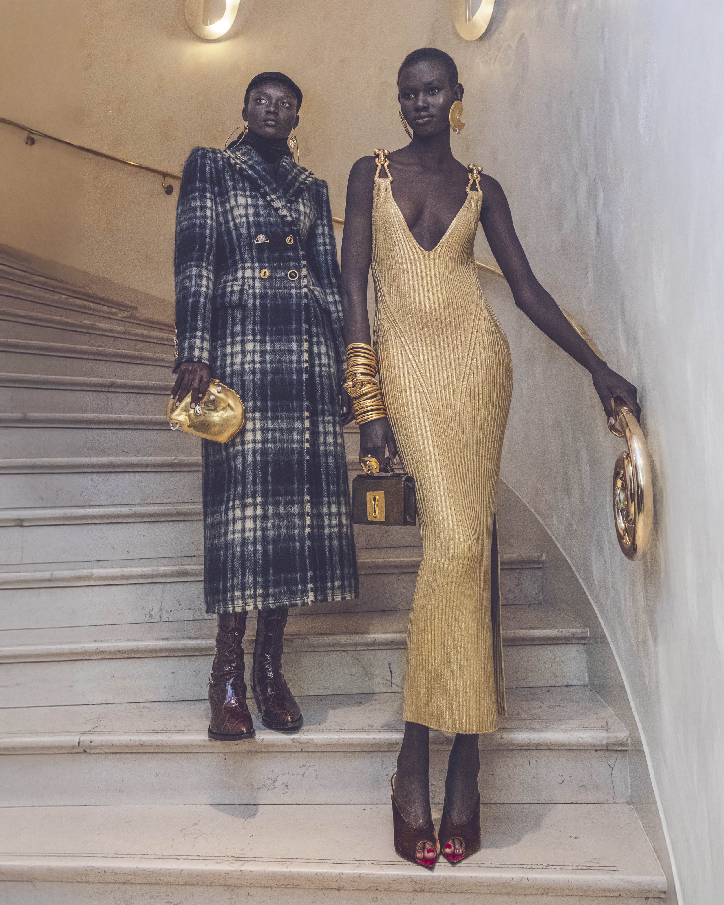 Two women with dark skin and short hair standing on a staircase, dressed in fashionable clothing and jewelry, looking serious. The woman on the left wears a checkered coat with gold buttons and holds a gold clutch. The woman on the right wears a gold