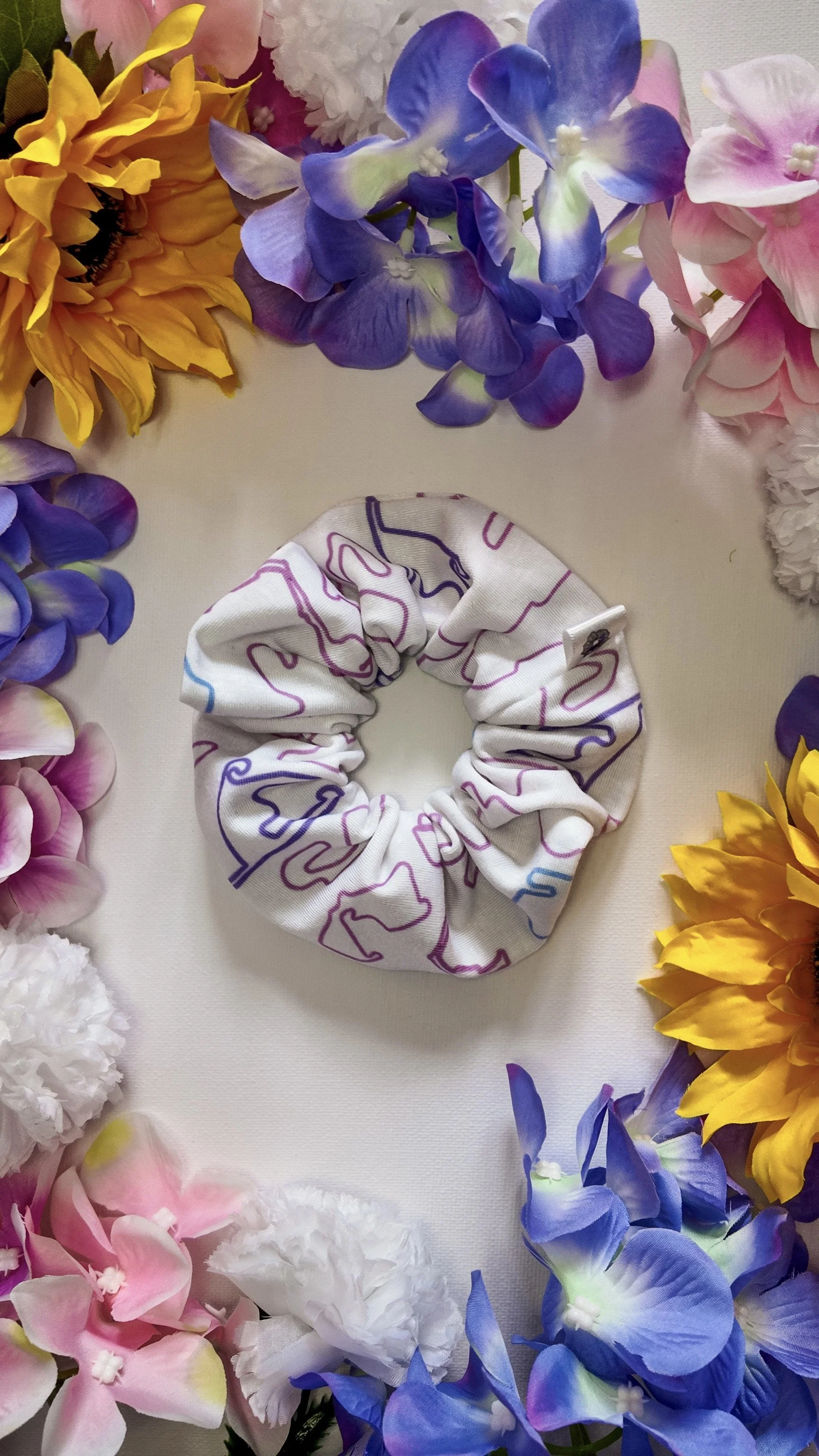 A fabric scrunchie with abstract, colorful line patterns in purple, pink, and blue on a white background, placed in the center of a circle of vibrant artificial flowers including sunflowers, orchids, and hydrangeas.