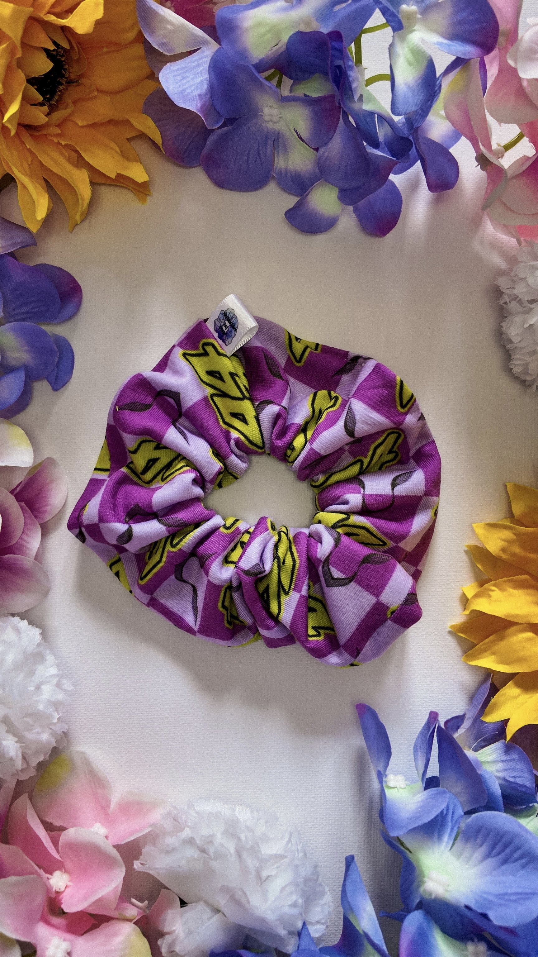A purple and yellow scrunchie with a repetitive cartoon logo pattern, placed on a white surface surrounded by an arrangement of colorful artificial flowers including sunflowers, pink, white, and purple flowers.