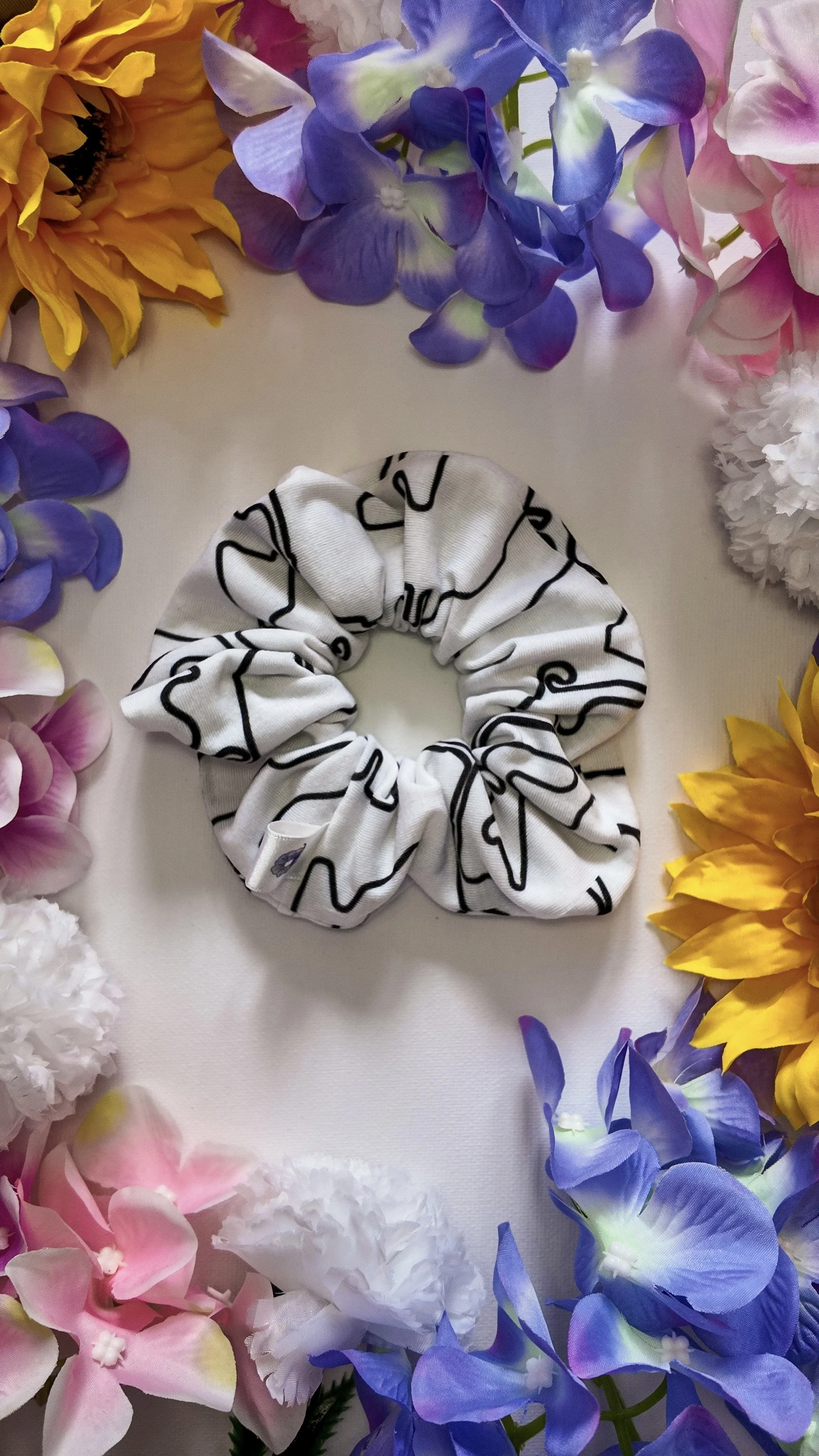 A black and white patterned fabric scrunchie surrounded by colorful artificial flowers, including pink, purple, white, and yellow blossoms, arranged in a circular pattern on a white background.