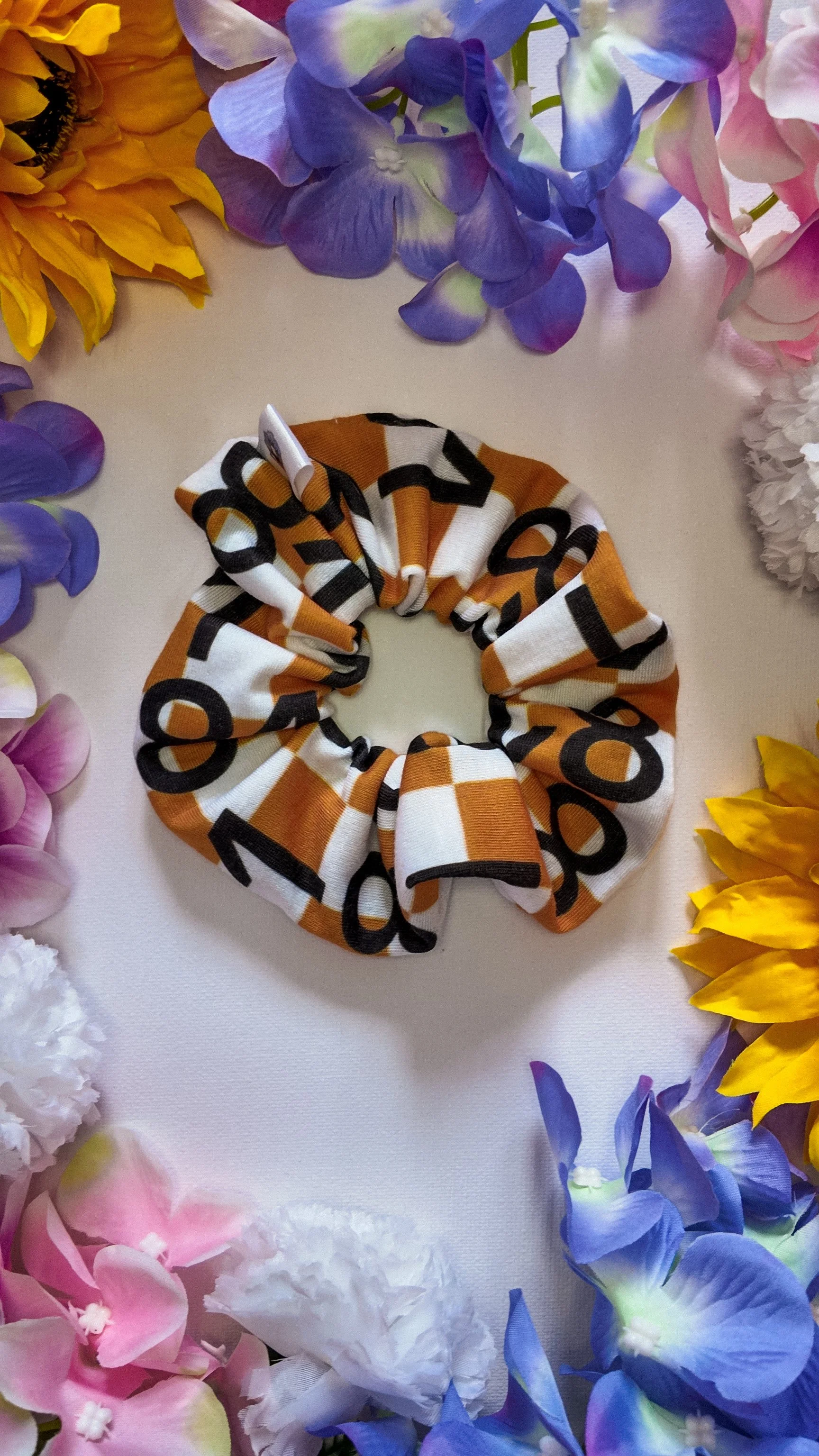 A scrunchie with orange, white, and black lettering design surrounded by colorful artificial flowers including purple, pink, yellow, and white flowers.