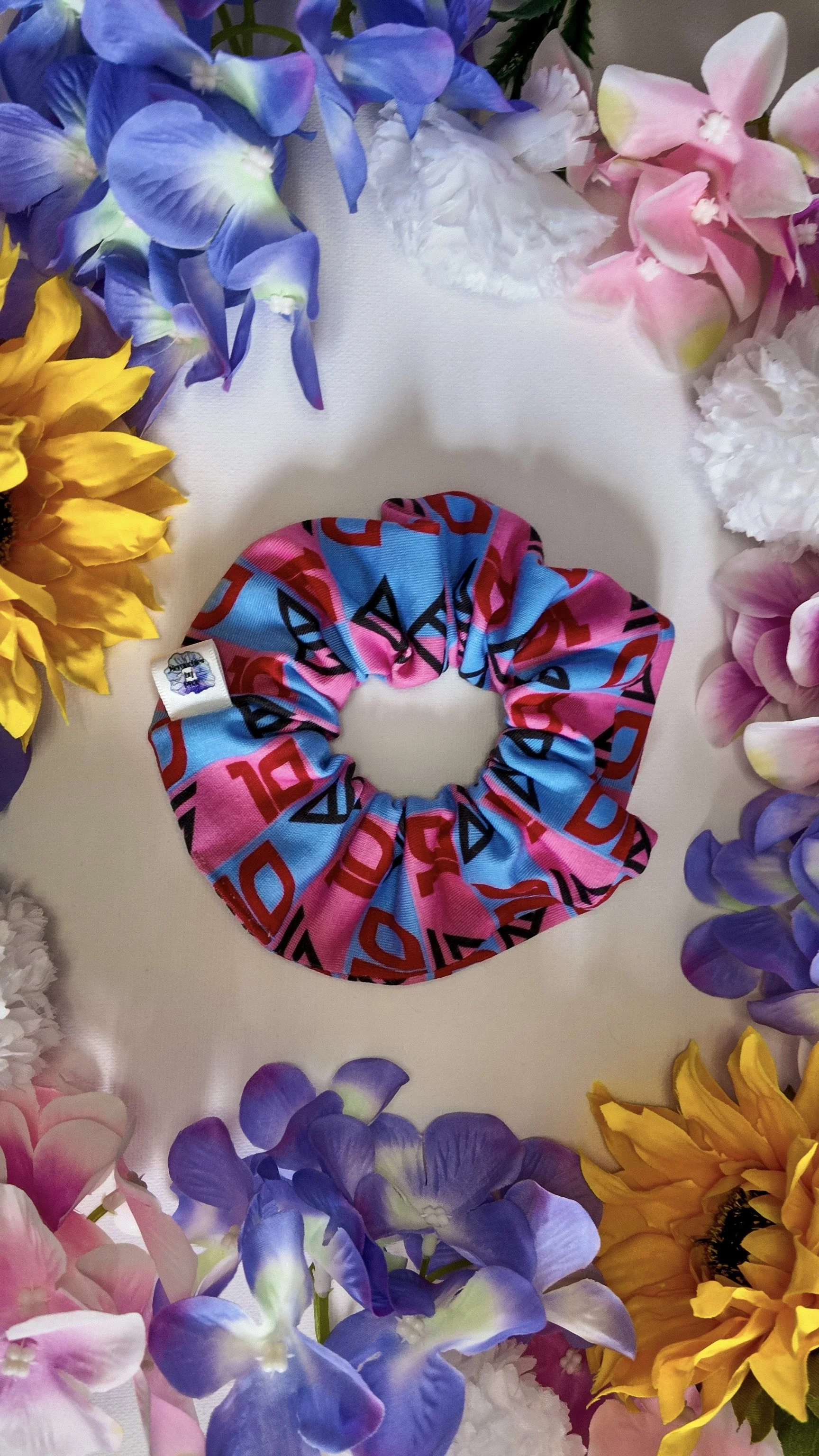 A colorful scrunchie with pink, blue, and red patterns is placed on a white surface, surrounded by a border of various artificial flowers including sunflowers, purple, pink, and white flowers.