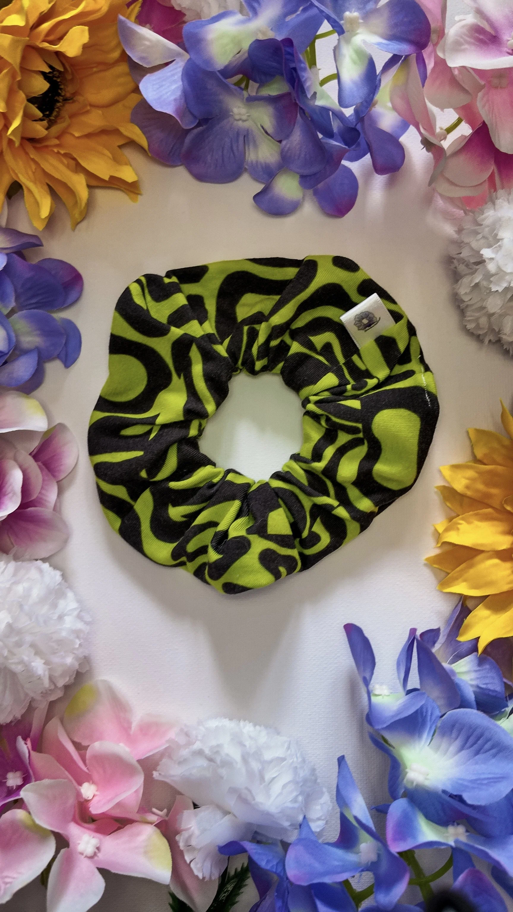 A black and neon green scrunchie with a bold, abstract pattern, surrounded by colorful artificial flowers, including sunflowers, hydrangeas, roses, and peonies, on a white surface.