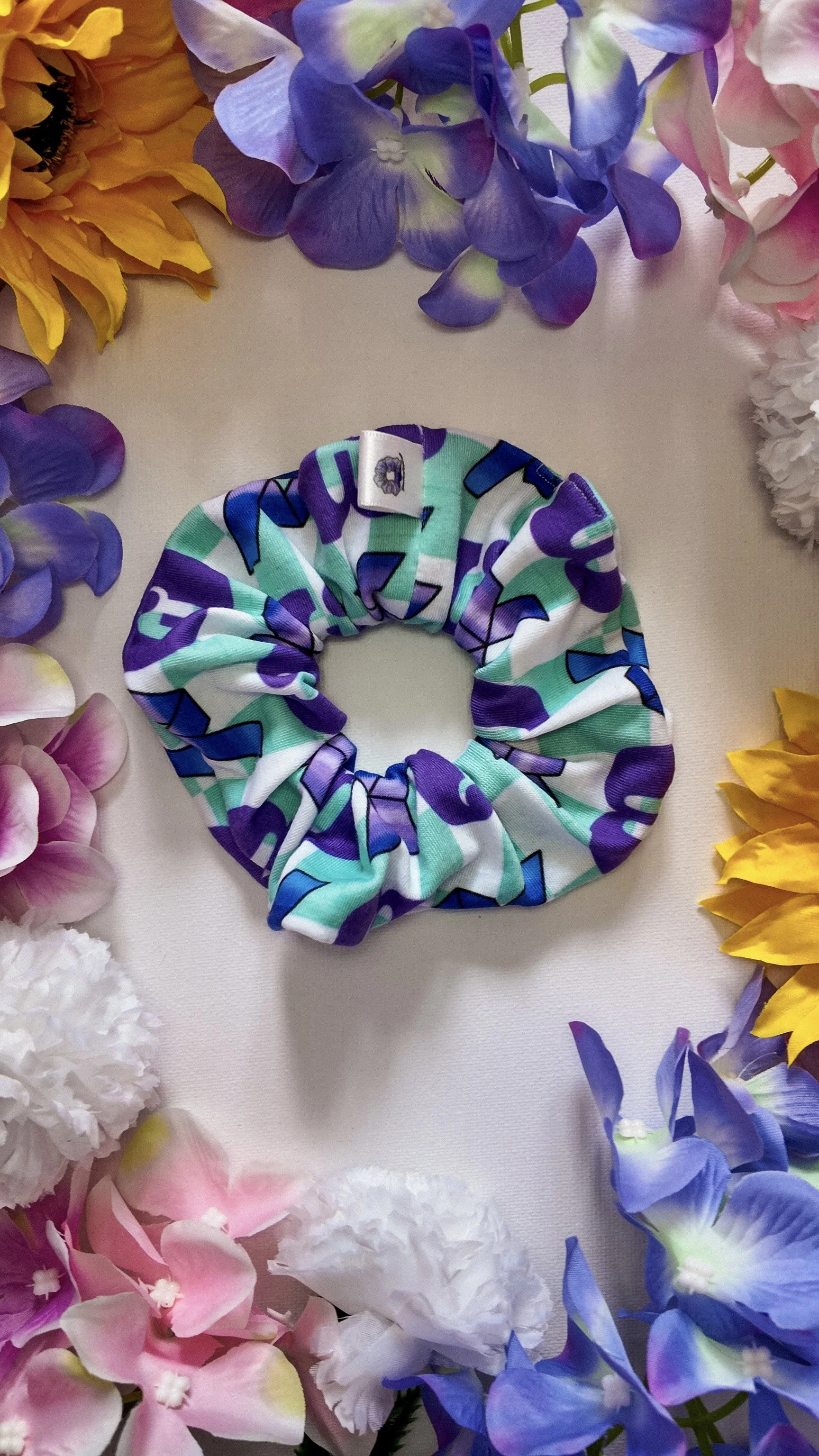 A colorful fabric scrunchie with a purple, blue, green, and white pattern lies on a white surface, surrounded by artificial flowers in purple, pink, white, and yellow.