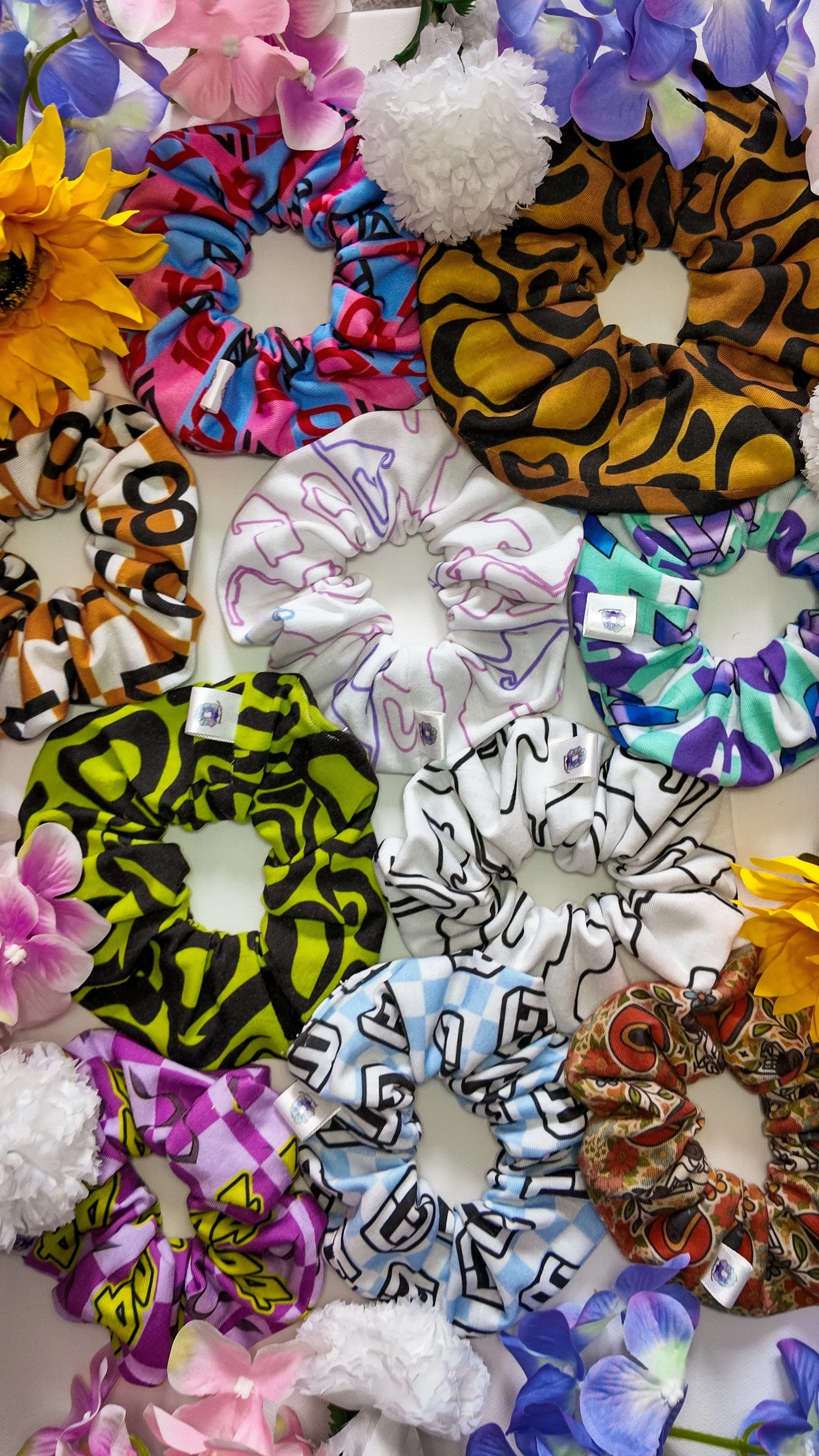 A variety of colorful fabric scrunchies arranged in a circular pattern, surrounded by pink, purple, yellow, white, and blue flowers.