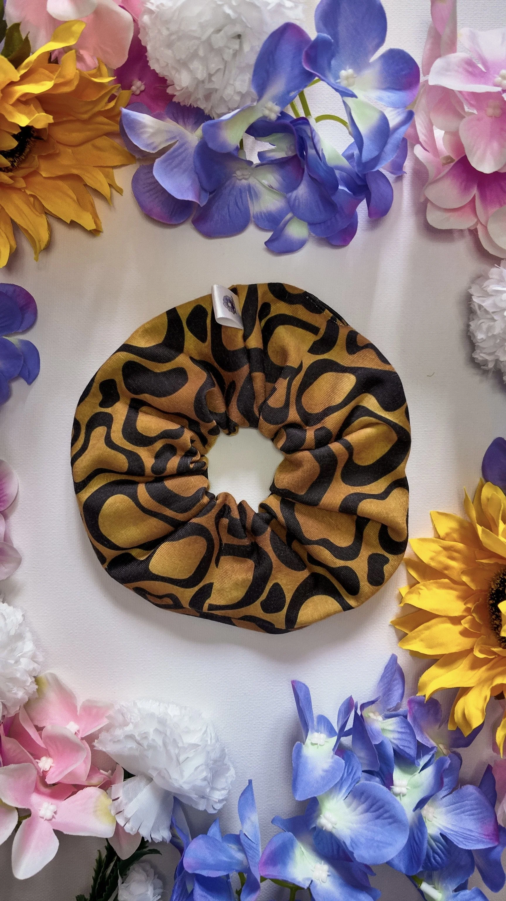 A leopard print scrunchie placed on a white surface surrounded by colorful artificial flowers, including sunflowers, pink, white, blue, and purple blooms.