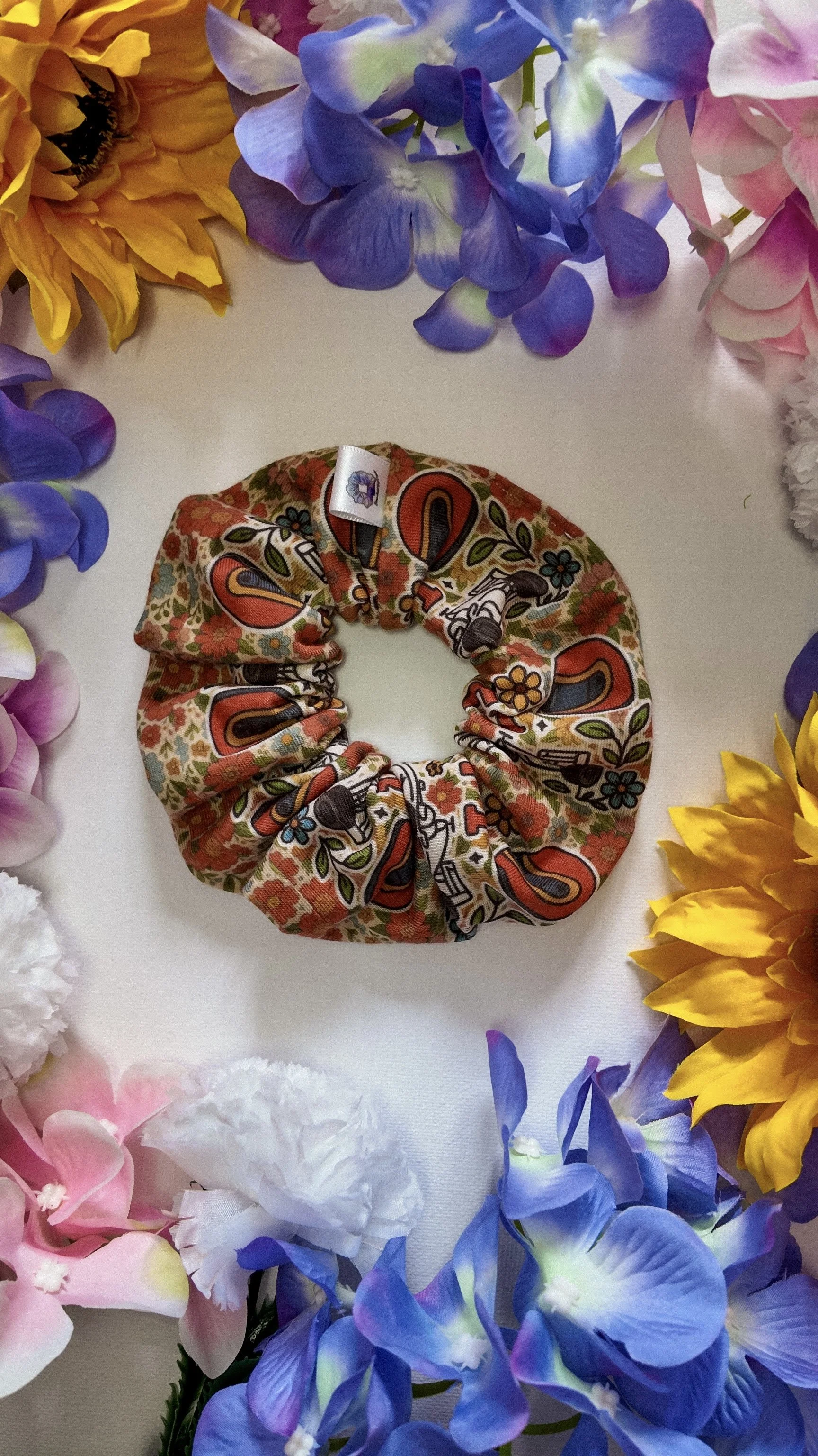 A colorful scrunchie with a paisley and floral pattern, placed on a white surface surrounded by an arrangement of various artificial flowers in purple, pink, yellow, blue, and white.