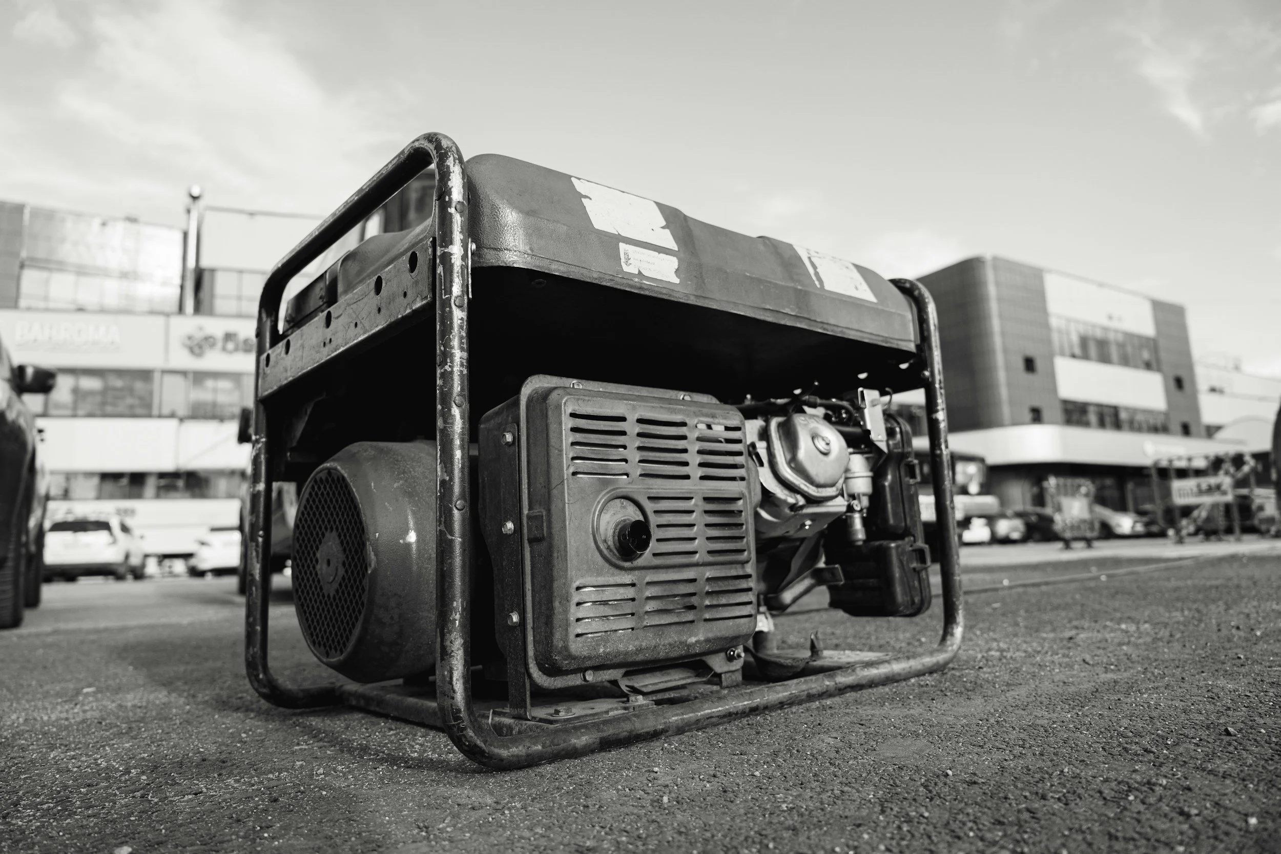 A close-up black and white photo of a portable generator on the ground in a parking lot with cars, buildings, and a cloudy sky in the background.
