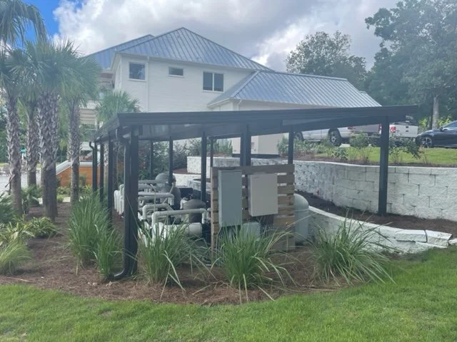 Carport structure with gas meters and piping underneath, surrounded by grass and plants, near a two-story house with a blue metal roof.