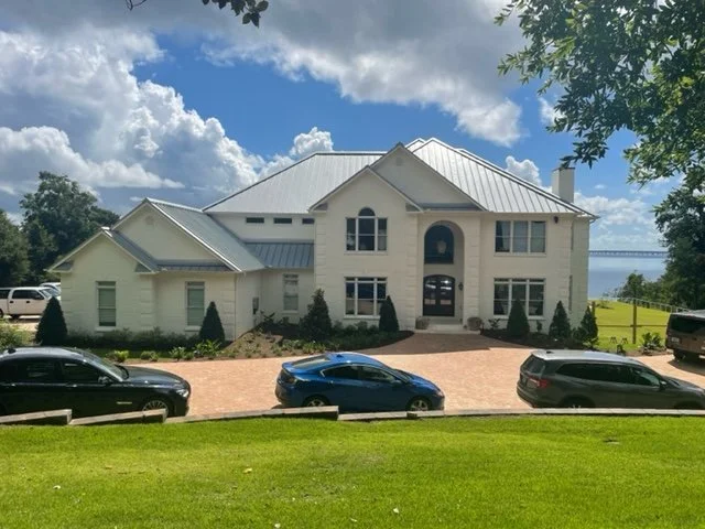 Large white house with a metal roof, surrounded by a driveway with three parked cars, green lawn, and trees, with water and cloudy sky in the background.