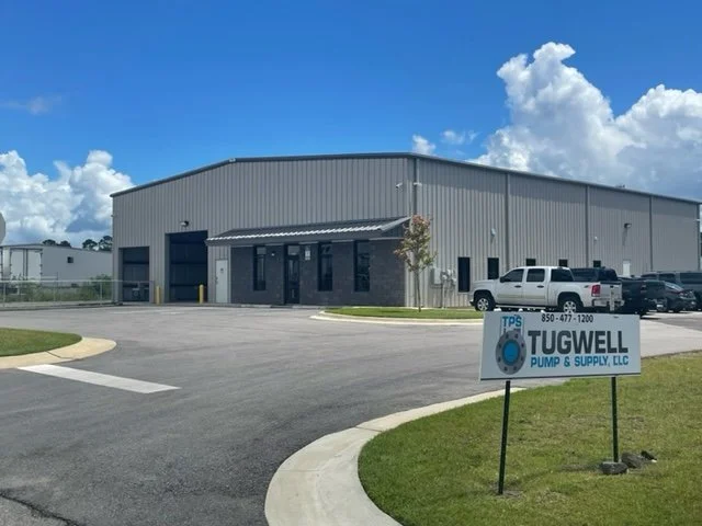 Industrial building with a sign for Tugwell Pump & Supply, LLC in front, parking lot with some trucks and cars, and partly cloudy sky.
