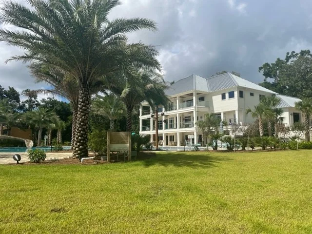 Large white modern beachfront house with multiple balconies, surrounded by palm trees and lush green lawn, under a cloudy sky.